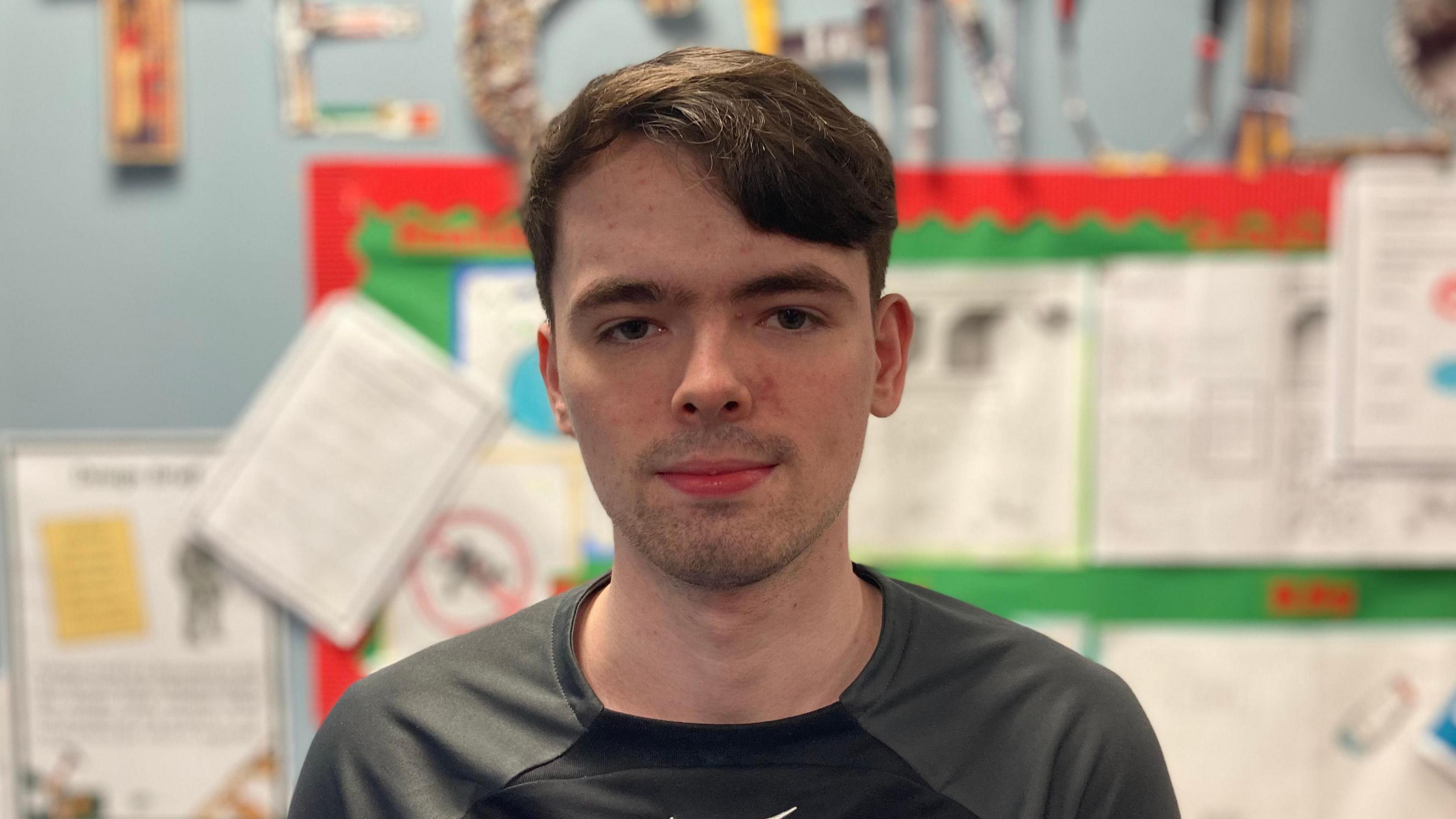 Joseph has short brown hair to one side and is wearing a grey t shirt. He is looking into the camera and behind him is a classroom display board. 