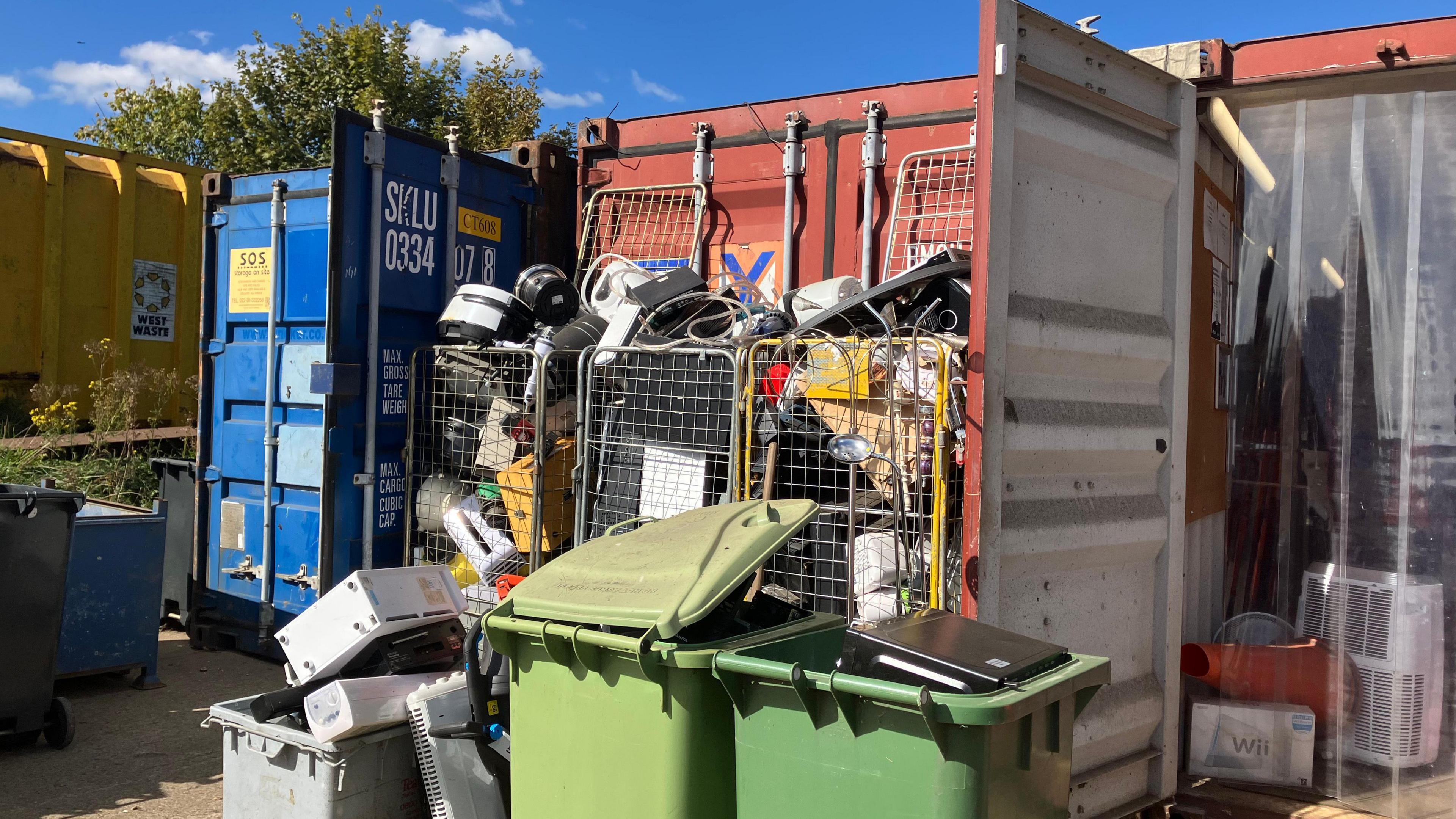 Two green bins, surrounded by three recycling crates and three trollies. They all have rubbish and recycling inside them.