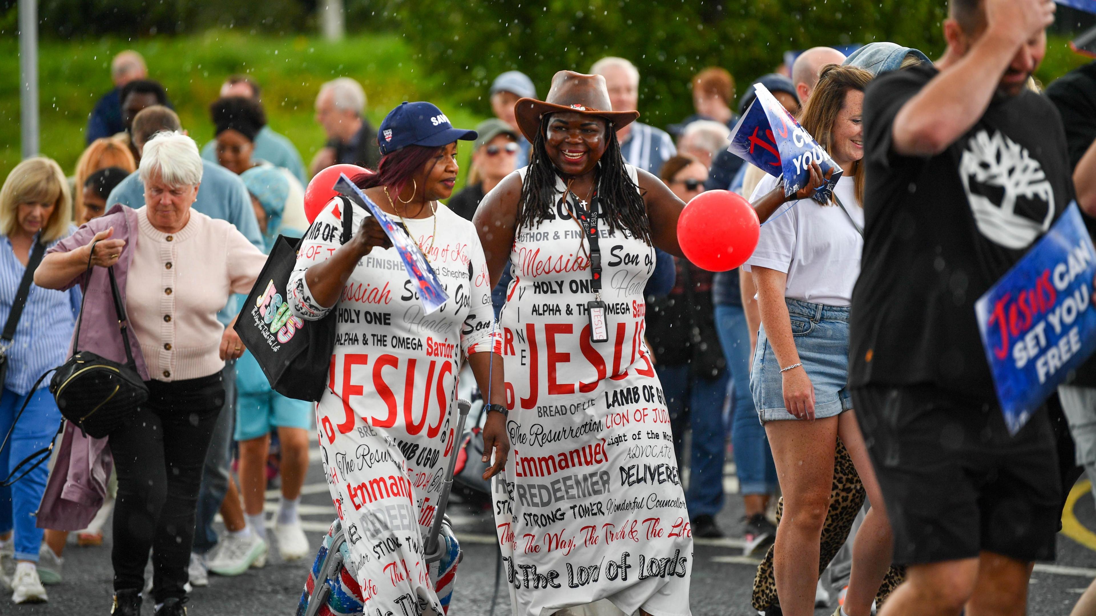 A group of people march down a road. Two ladies are dressed in white dresses which say words like Jesus and Redeemer on it.