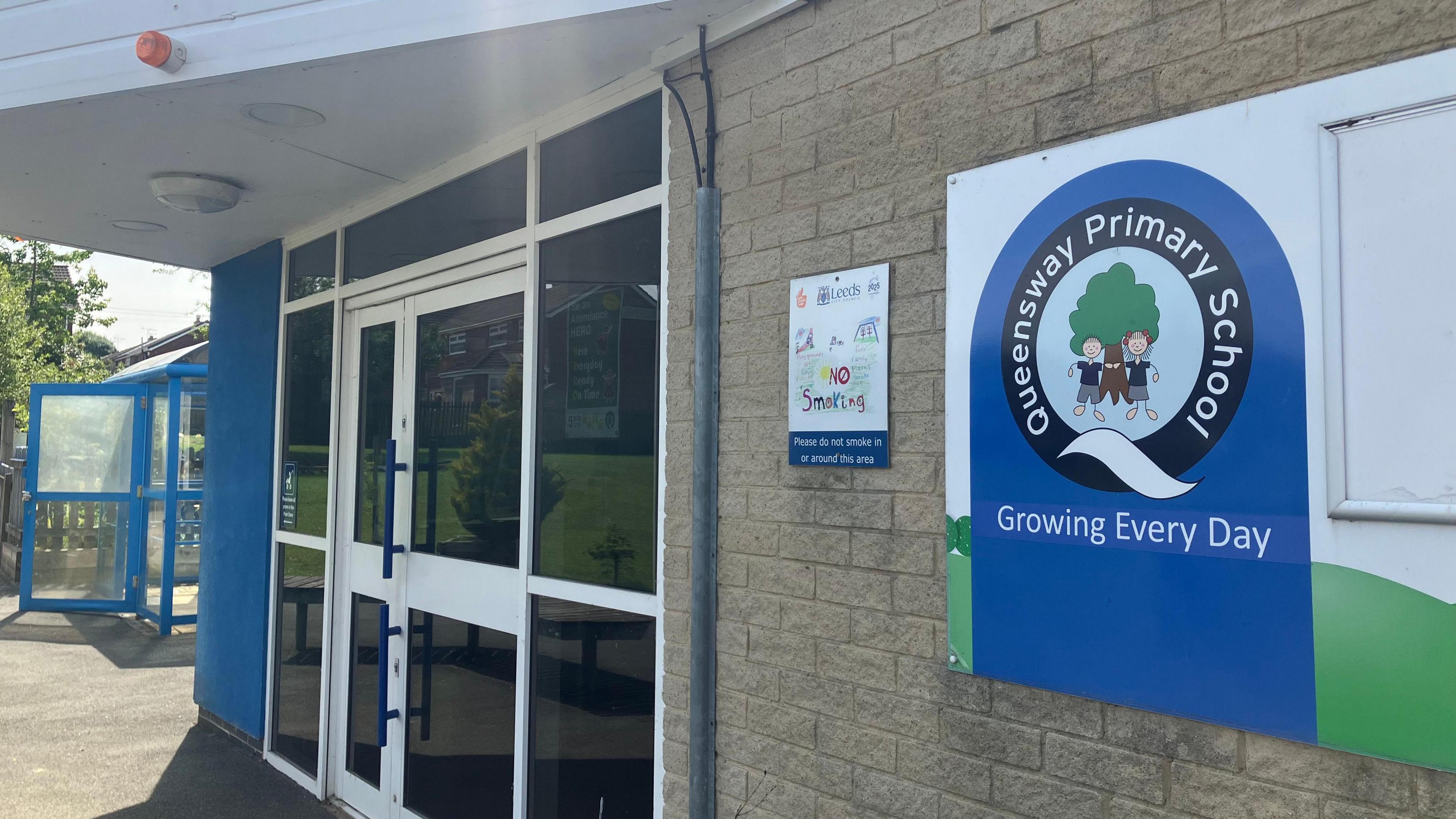 The front door of Queensway Primary School. There is a door on the left, with a blue and green sign on a brick wall on the right hand side.