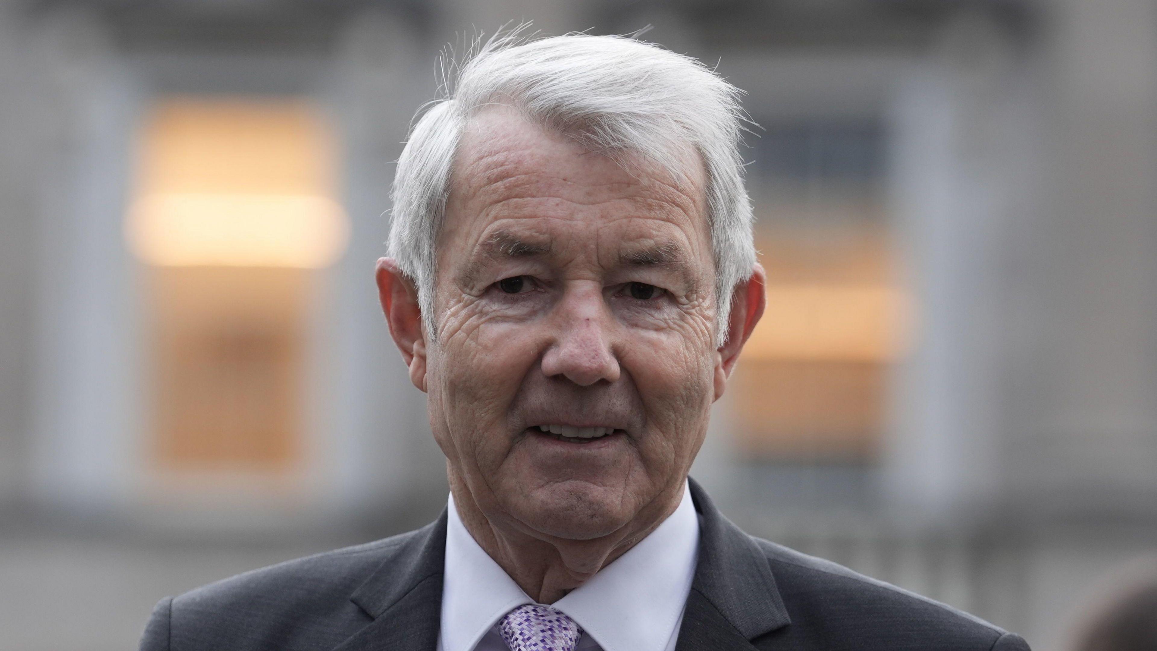 A man with grey hair, pictured from the shoulders up, wearing a grey suit and lilac shirt. The background of the image is blurred. 