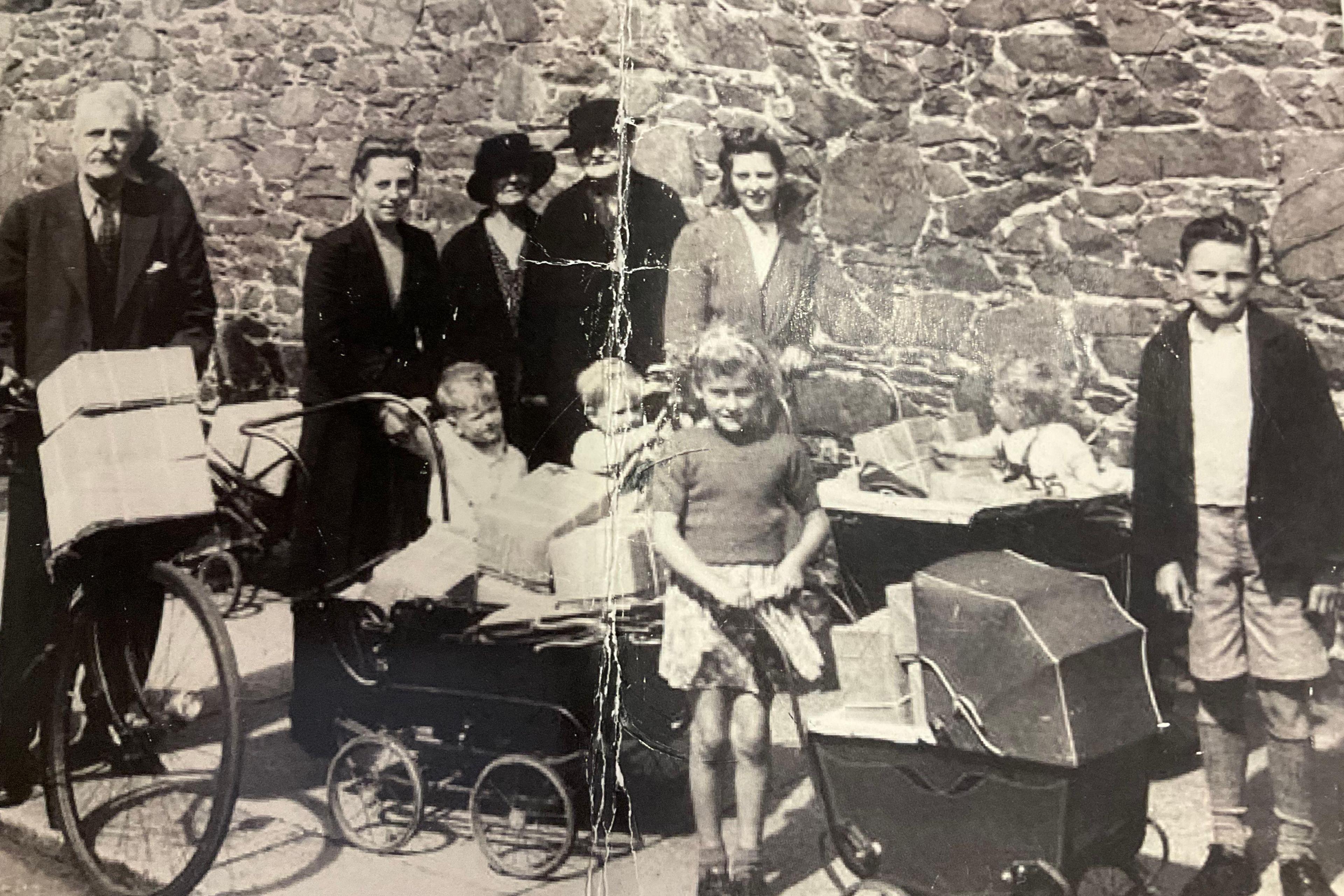 A black and white World War Two era photo of a family carrying packages wrapped with paper and string - standing in front of a granite wall. There are two older adult men - one on a bicycle with packages on the front. Next to them three adult women and three toddlers, a young girl holding a pram full of packages, and a young boy in shorts,