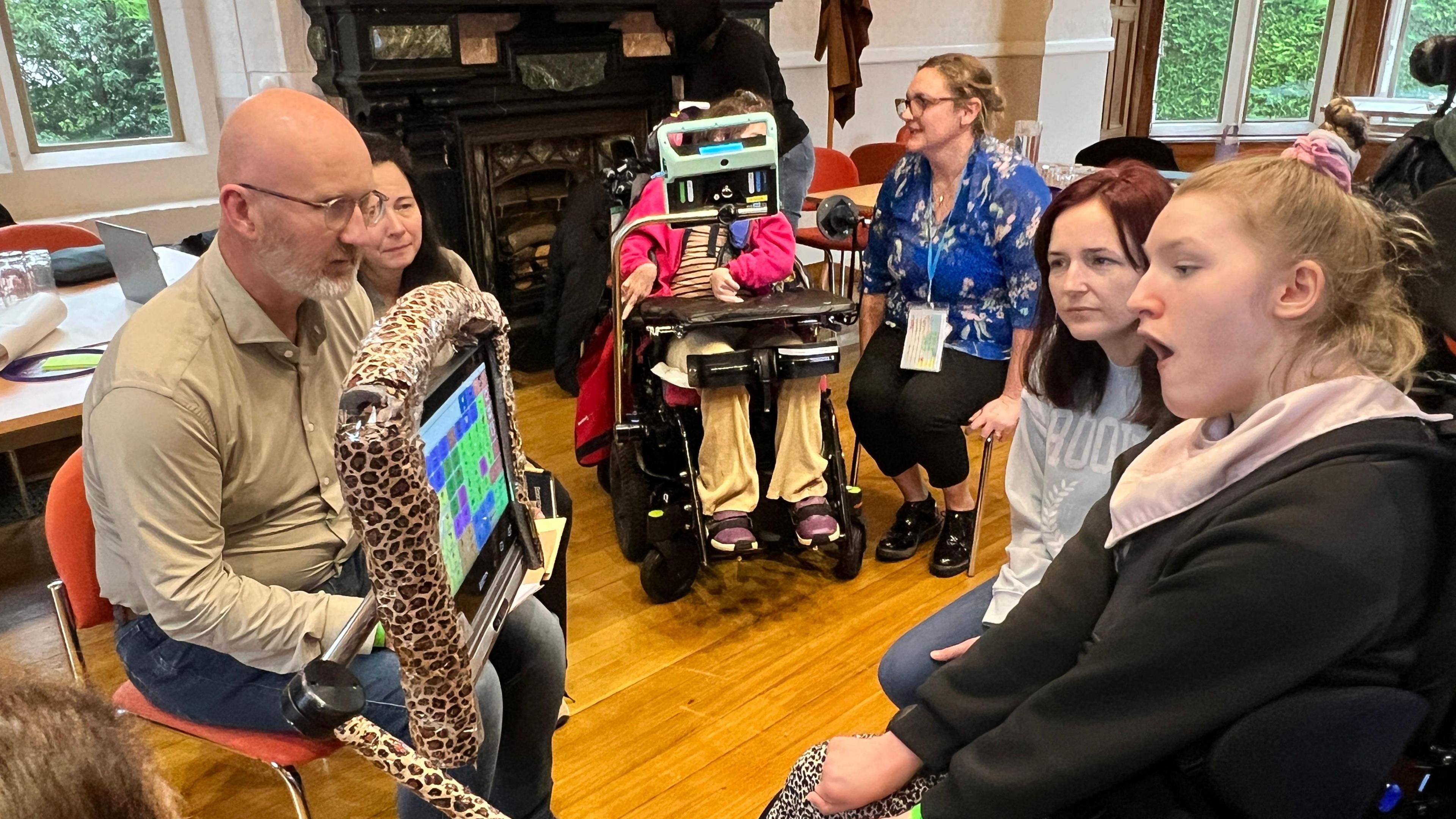 A group of people in a room with wooden flooring and large windows. Two individuals in wheelchairs are using assistive communication devices, including a screen with a leopard-print frame. Others are seated on orange chairs, and a dark ornate fireplace is visible in the background.