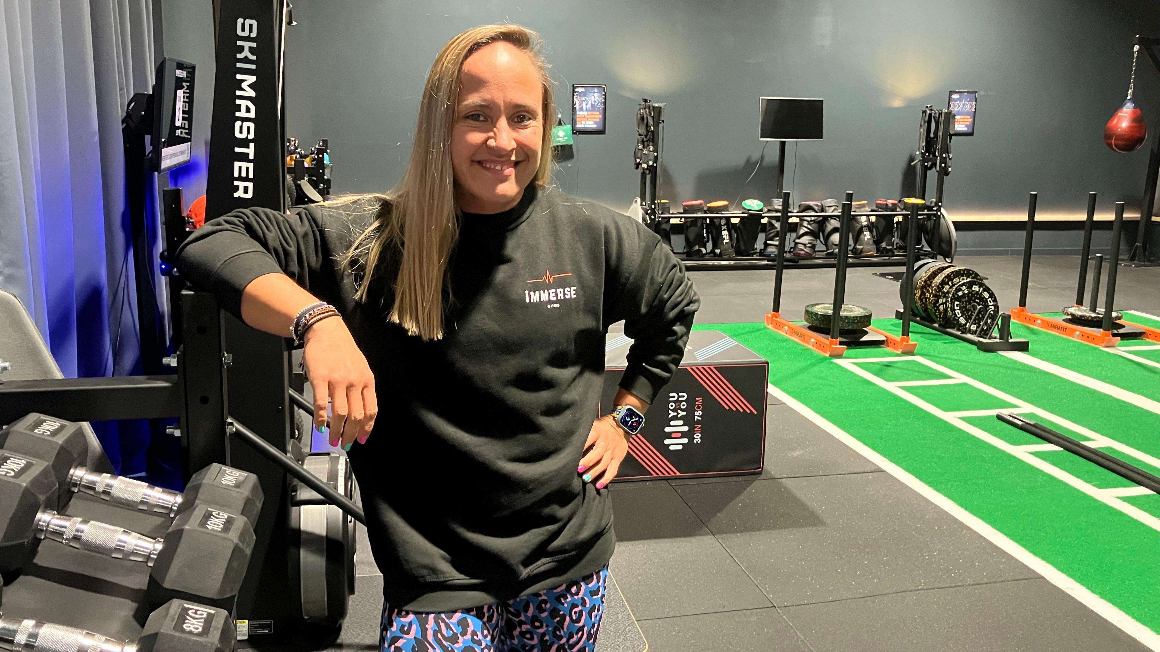 Sophia Gaiqui leaning on some dumbbells. There are free weights in the background and some exercise ropes. 