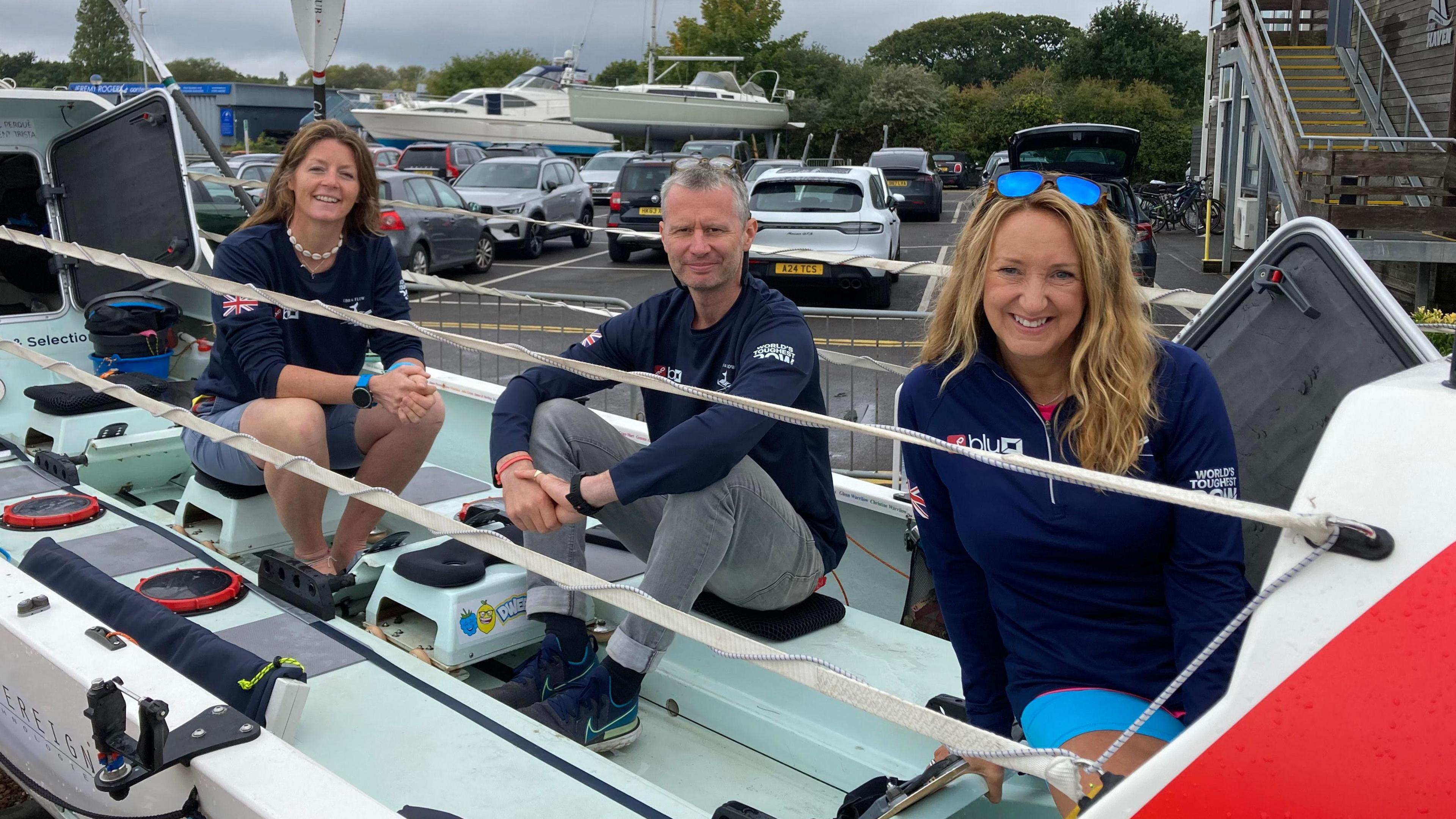 Two women and a man sat on a rowing boat on land staring at the camera and smiling