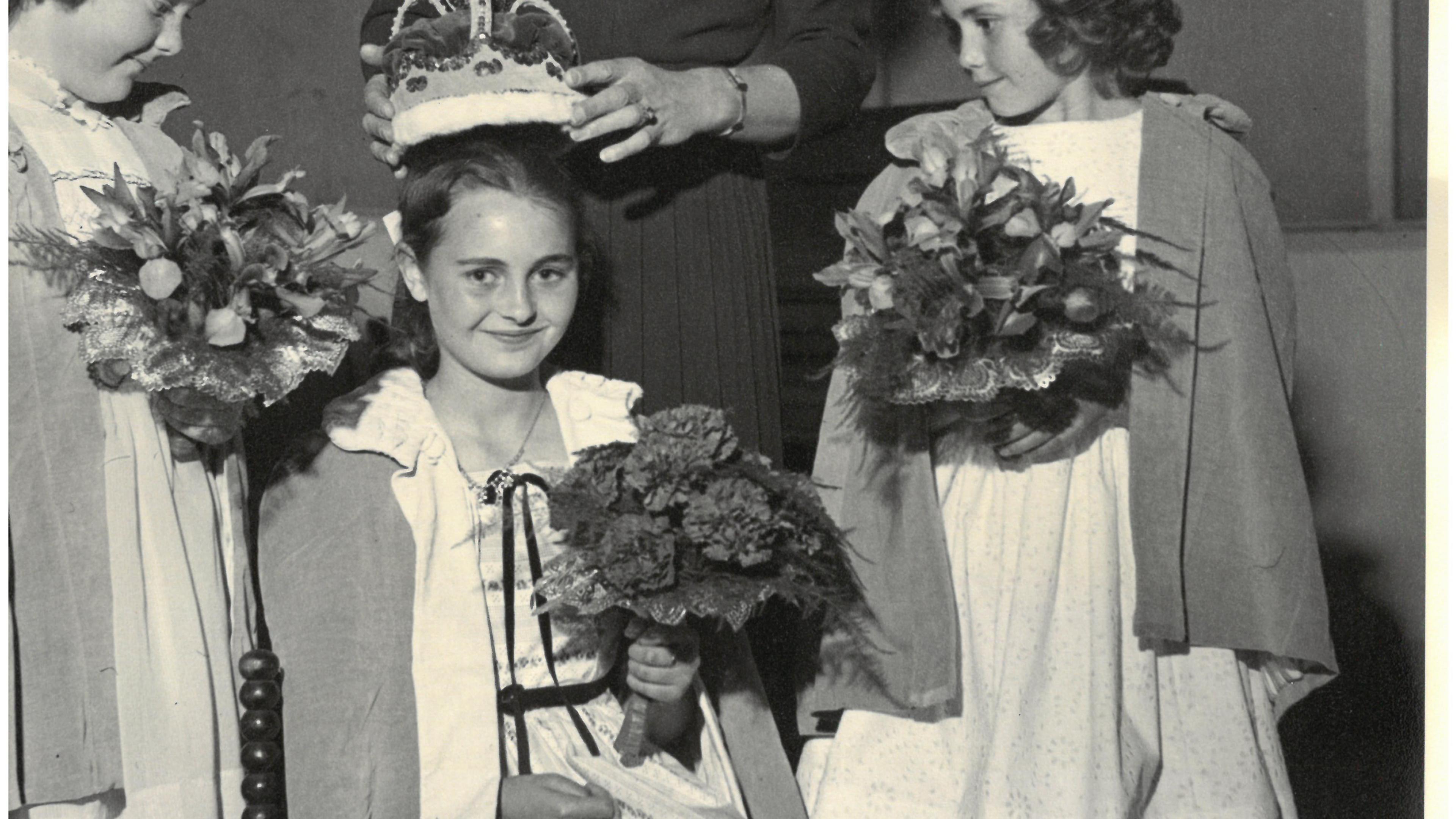 Black and white image. A young girl sits in a chair as a crown is placed on her head. She is holding a bunch of flowers. Two young girls stand beside her, holding flowers.