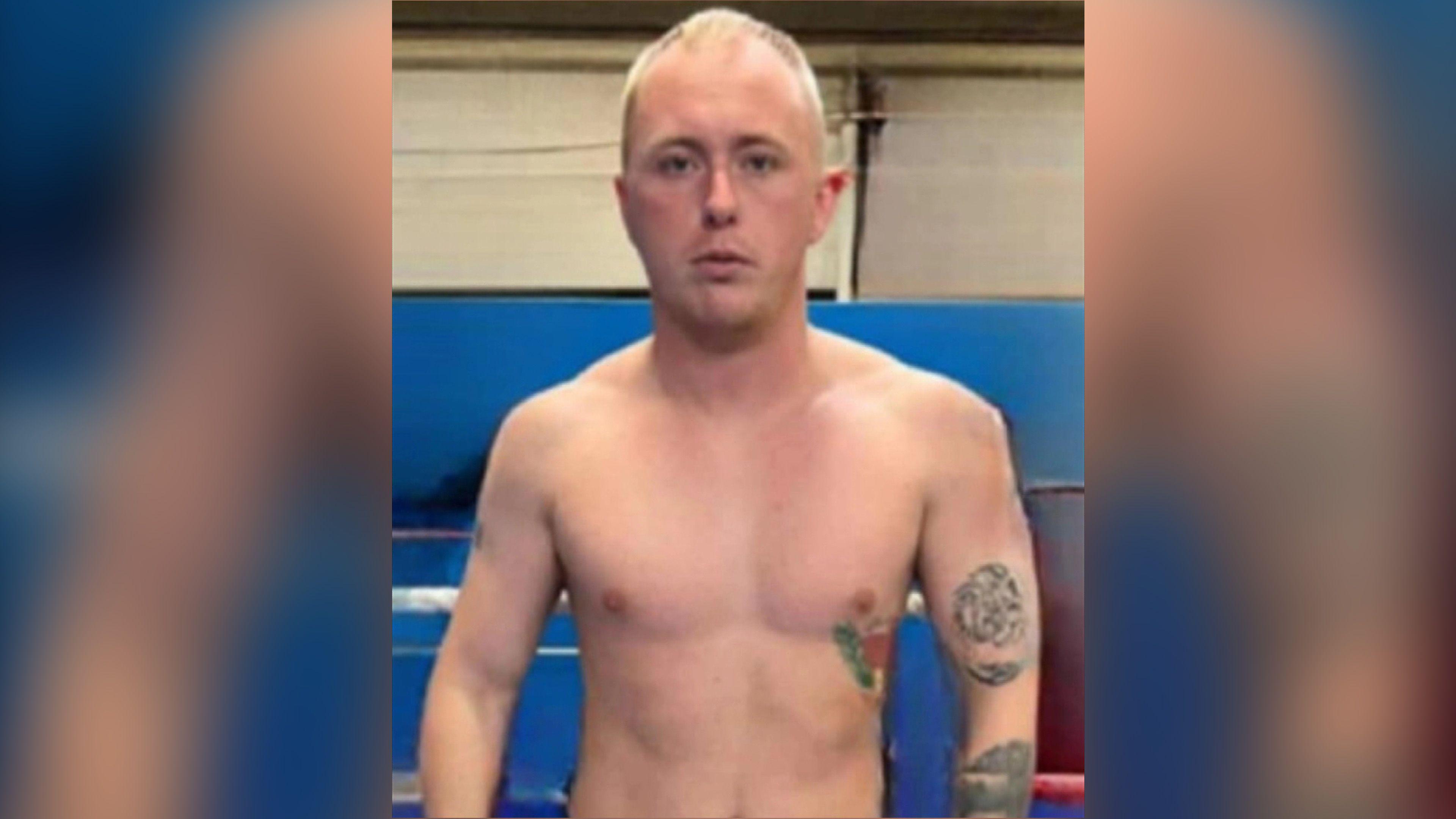 A man is standing in a fighting gym with no top on. He has short sandy hair and is looking straight at the camera. The man has several tattoos on his left arm. Behind him are the ropes of a boxing gym and a blue wall. 