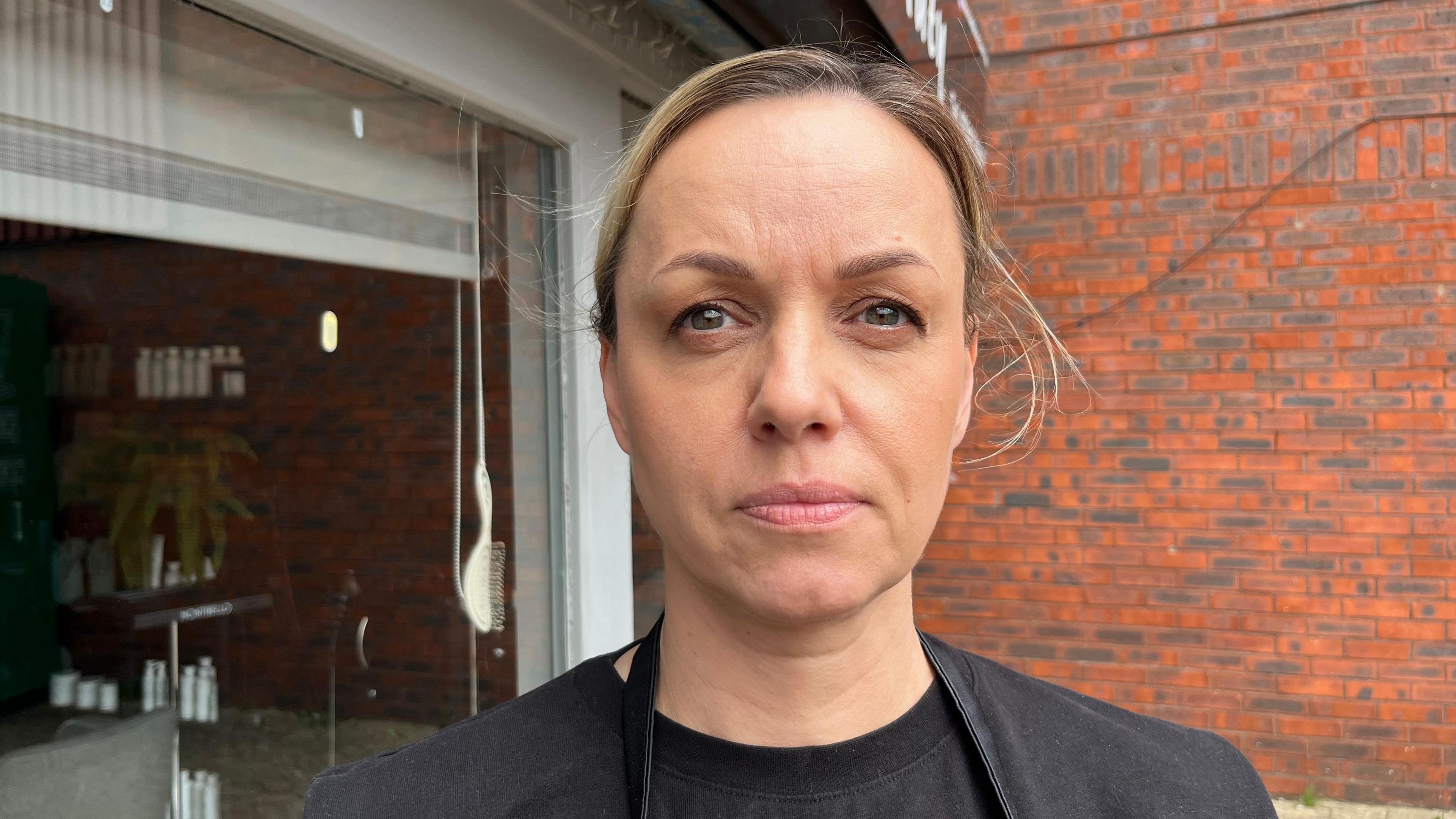 Jurga Ziliene is looking towards the camera and slightly frowning, wearing a black t-shirt. Behind her is a brick wall and an open garage door.