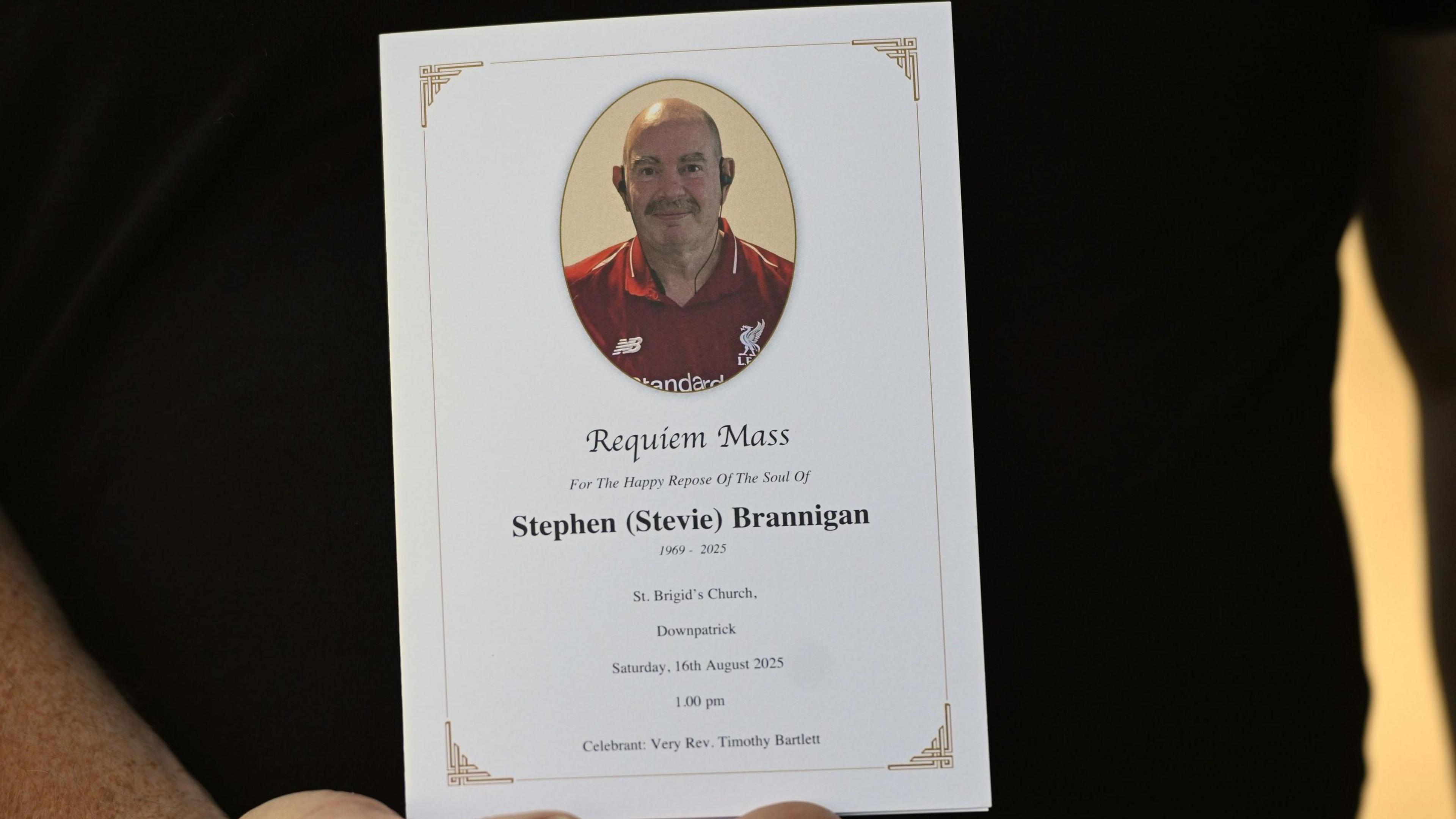 A funeral programme for Stephen 'Stevie' Brannigan is held up by a man in a back t-shirt. Mr Brannigan is bald with a dark moustache and is wearing a Liverpool top in the picture