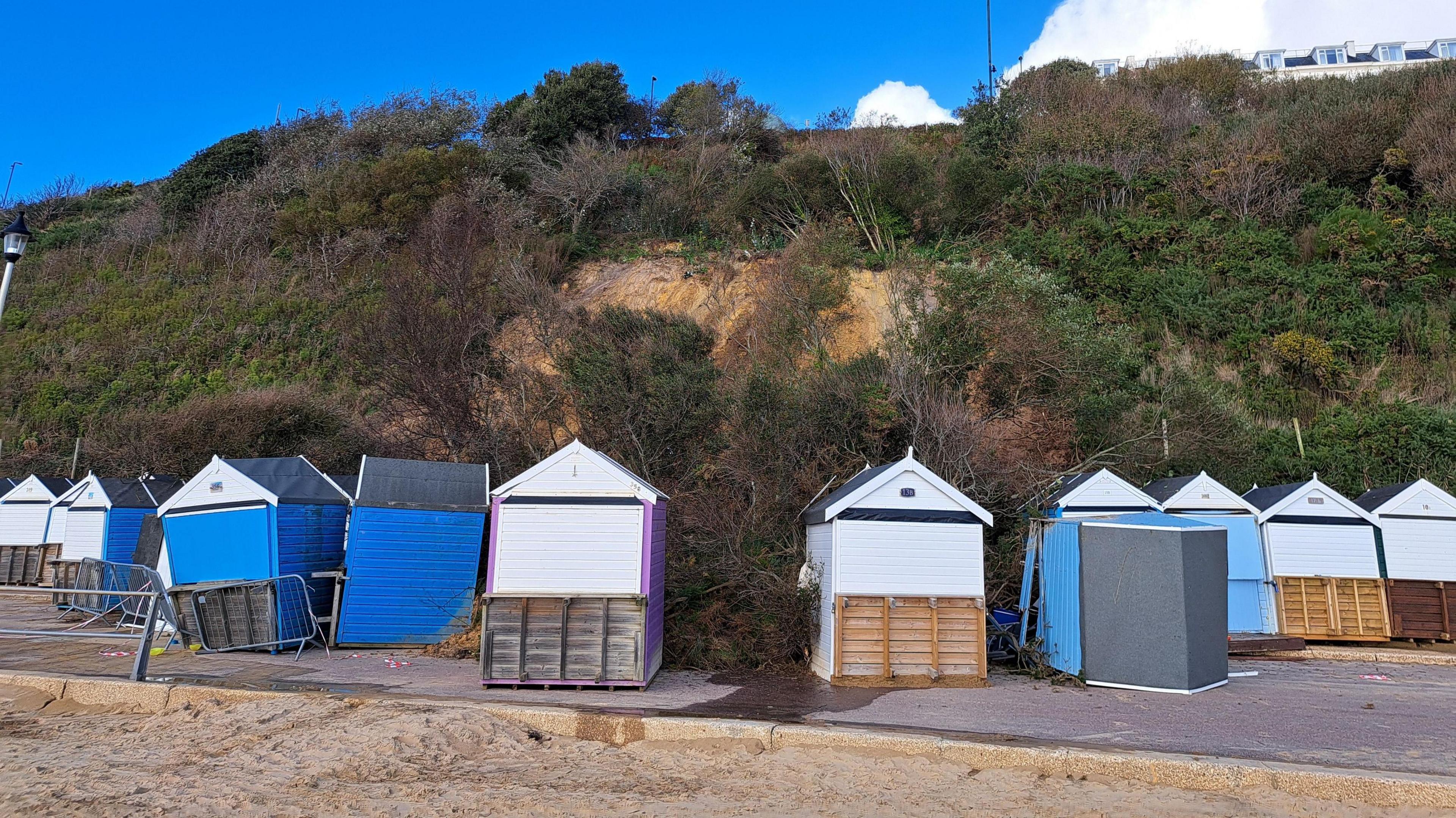 View of the huts impacted by the cliff slide from a different angle. Buildings can be seen on top of the cliff. It's a cloudy day.