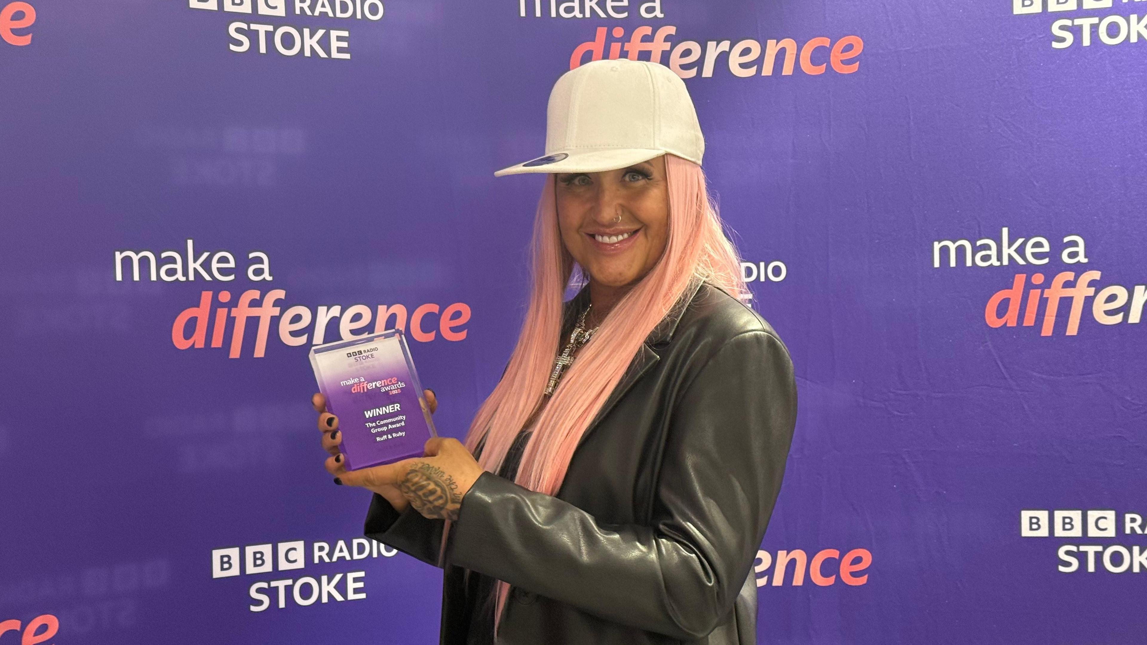 Dawnie Reynolds, a woman with pink hair is wearing a cream-coloured baseball cap and a black jacket. She is standing in front of a purple BBC Radio Stoke Make A Difference step and repeat board.