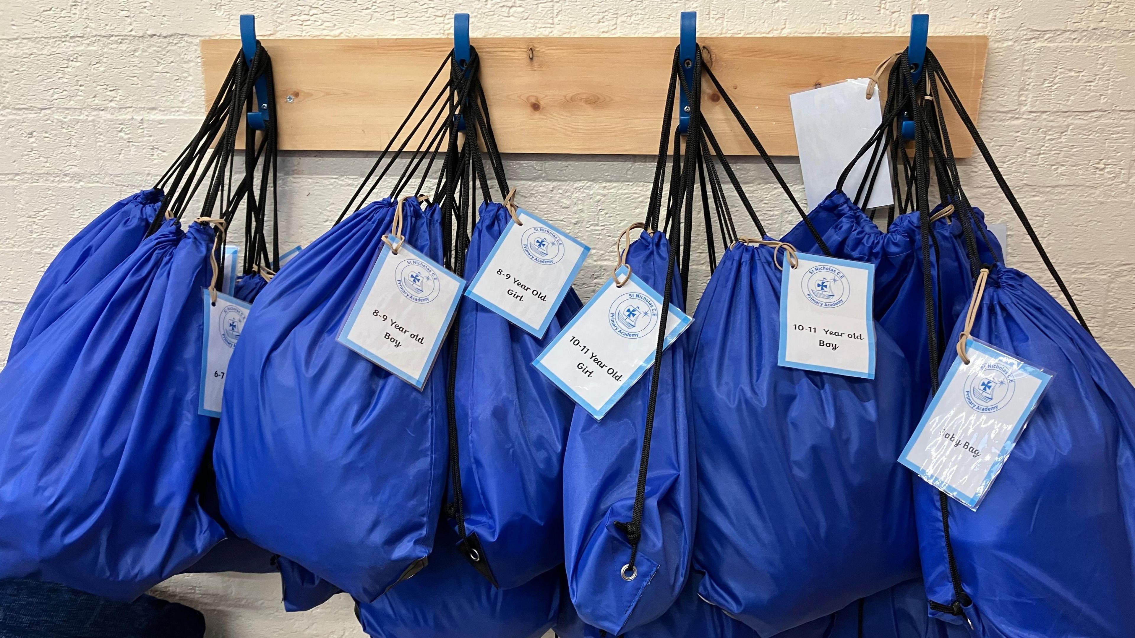 Ten blue draw-string bags hang from four wall pegs. Each bag is labelled with the age and gender of the child it has been packed for. 