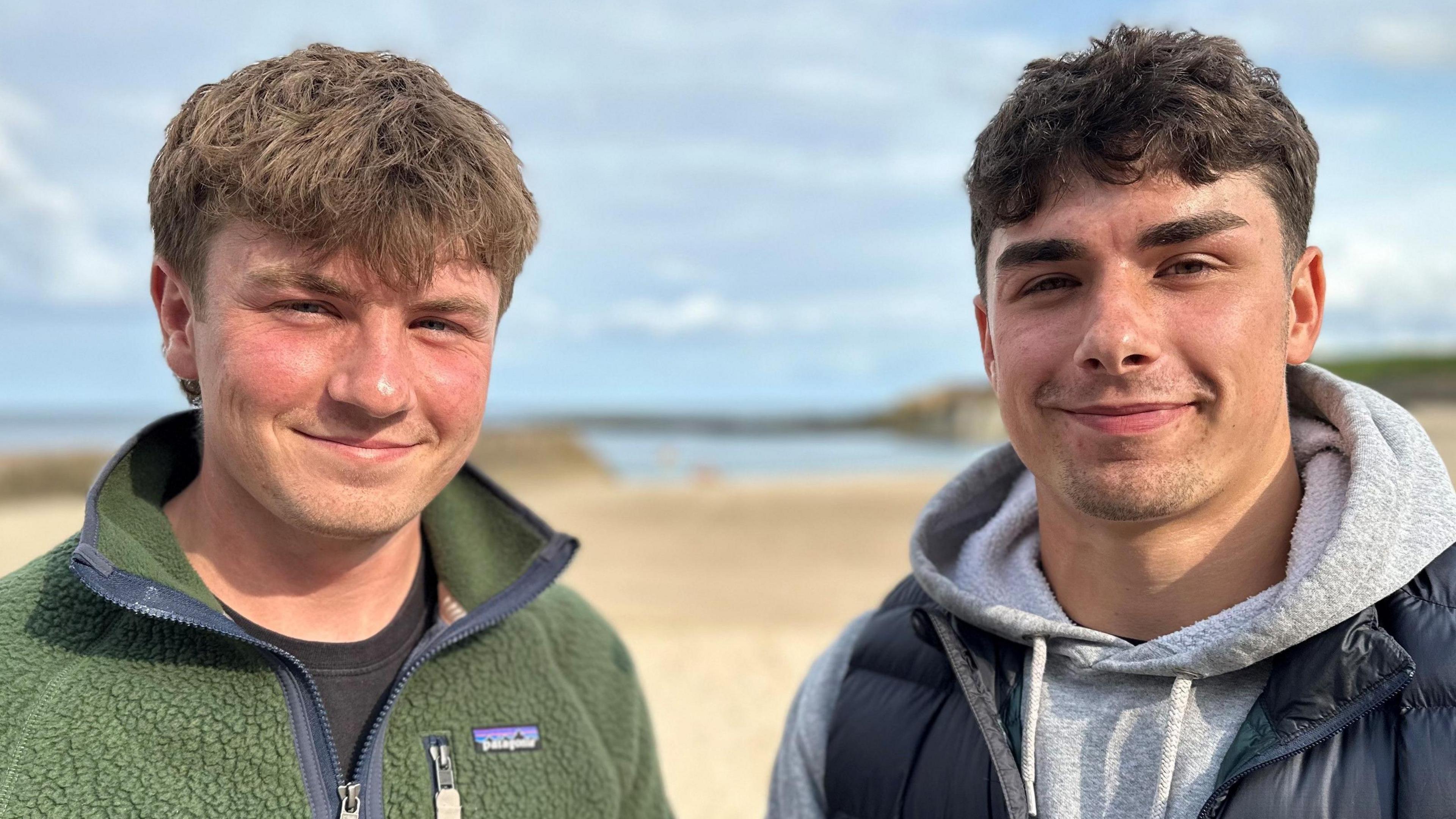 Euan and Andrew stand side by side smiling at the camera, with a yellow beach and blue sea behind them. Euan has brown hair and is wearing a green fleece. Andrew has black hair and is wearing a grey hoody beneath a navy body warmer.