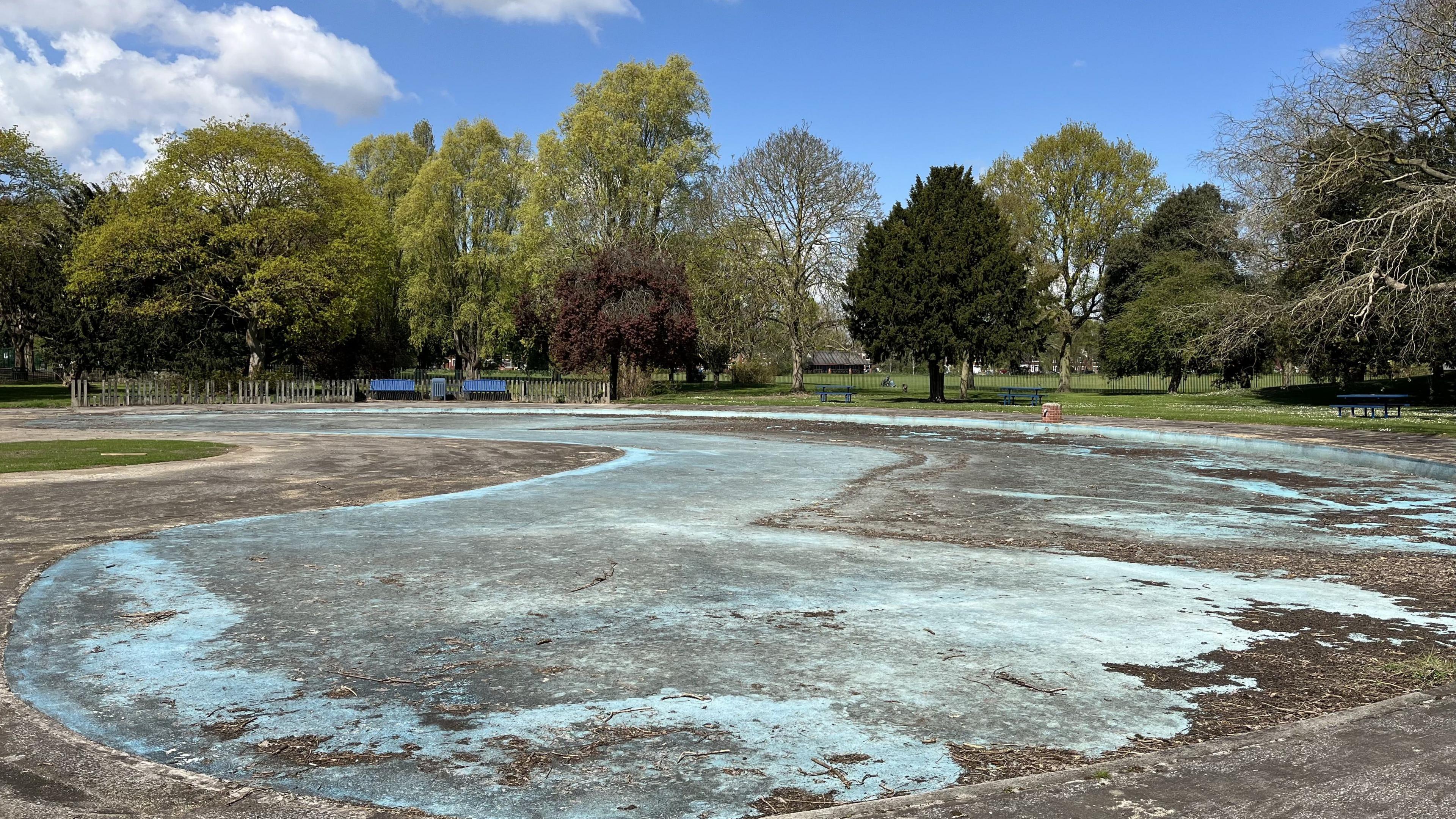 The closed paddling pool at Pickering Park in Hull. The blue-painted concrete is covered with leaves and dirt and surrounded by a large grassed area and trees. The sun is shining on the area with blue skies and cloud.