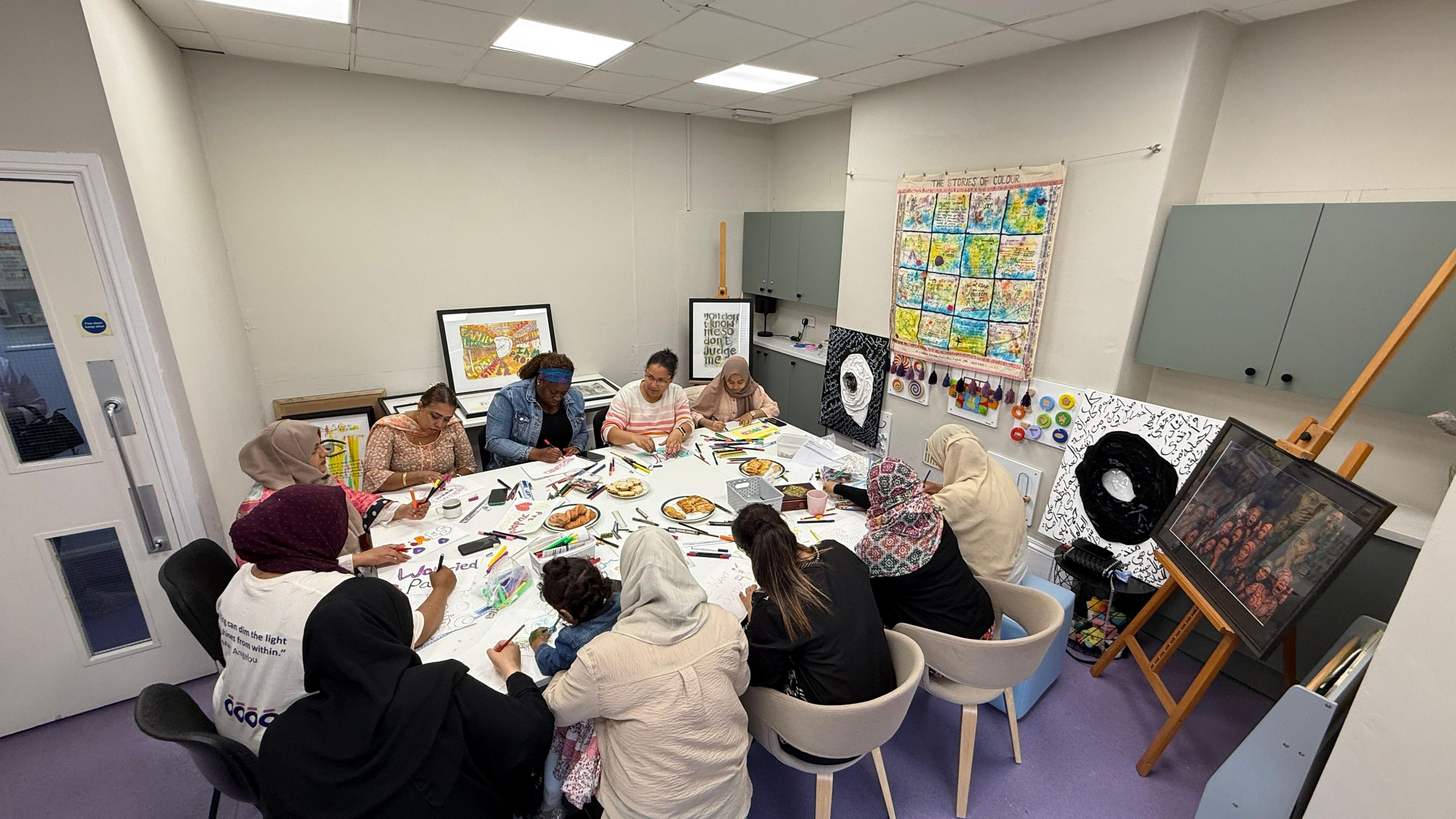 A group of women sit around a large table drawing and colouring pictures. Some are wearing hijabs and there are pieces of colourful art displayed on the walls around them. One woman has a young girl sitting on her lap.