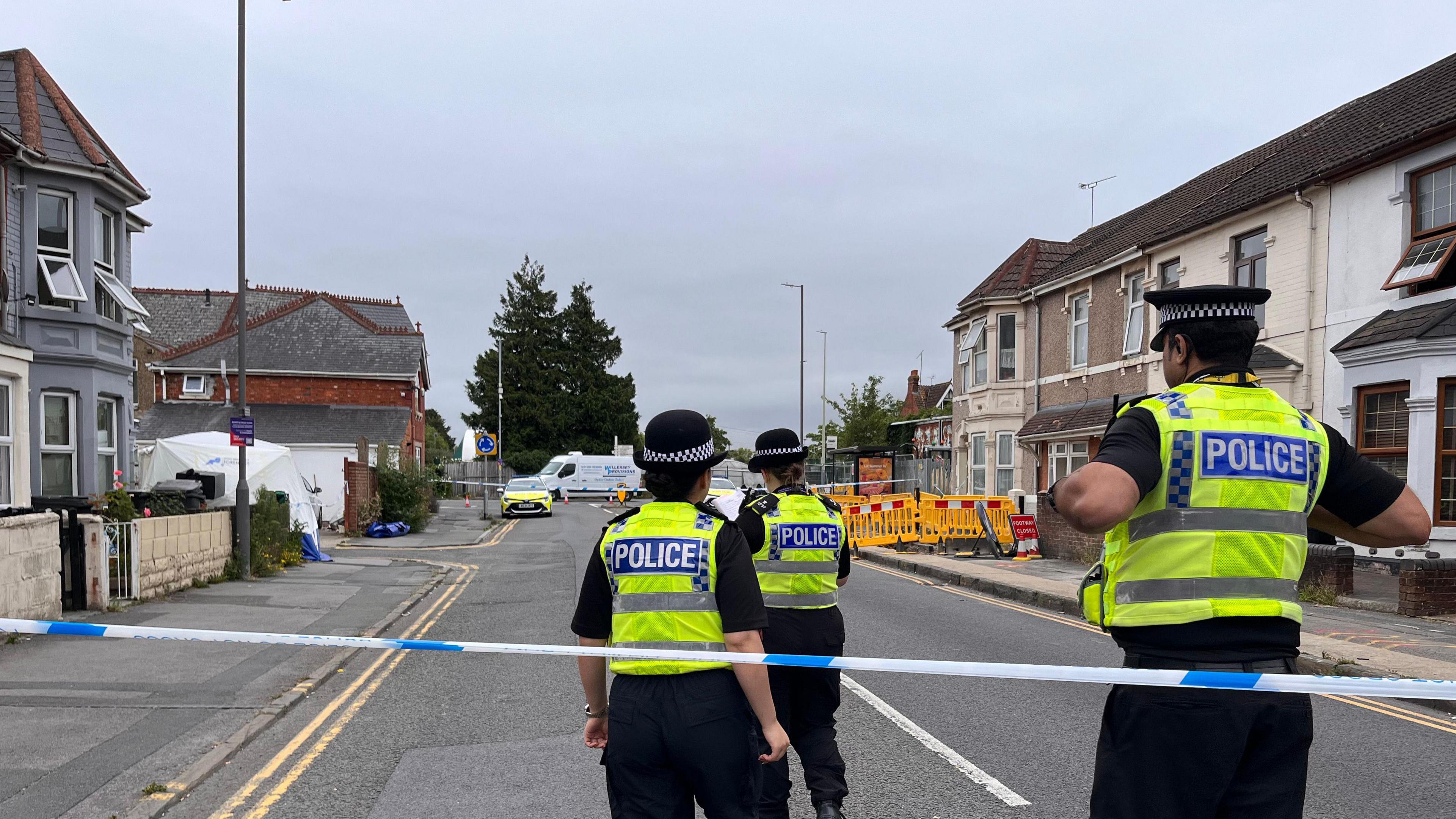Three police officers stood by police tape inside a police cordon. They are wearing high vis jackets and black police hats. You can see houses in the background.