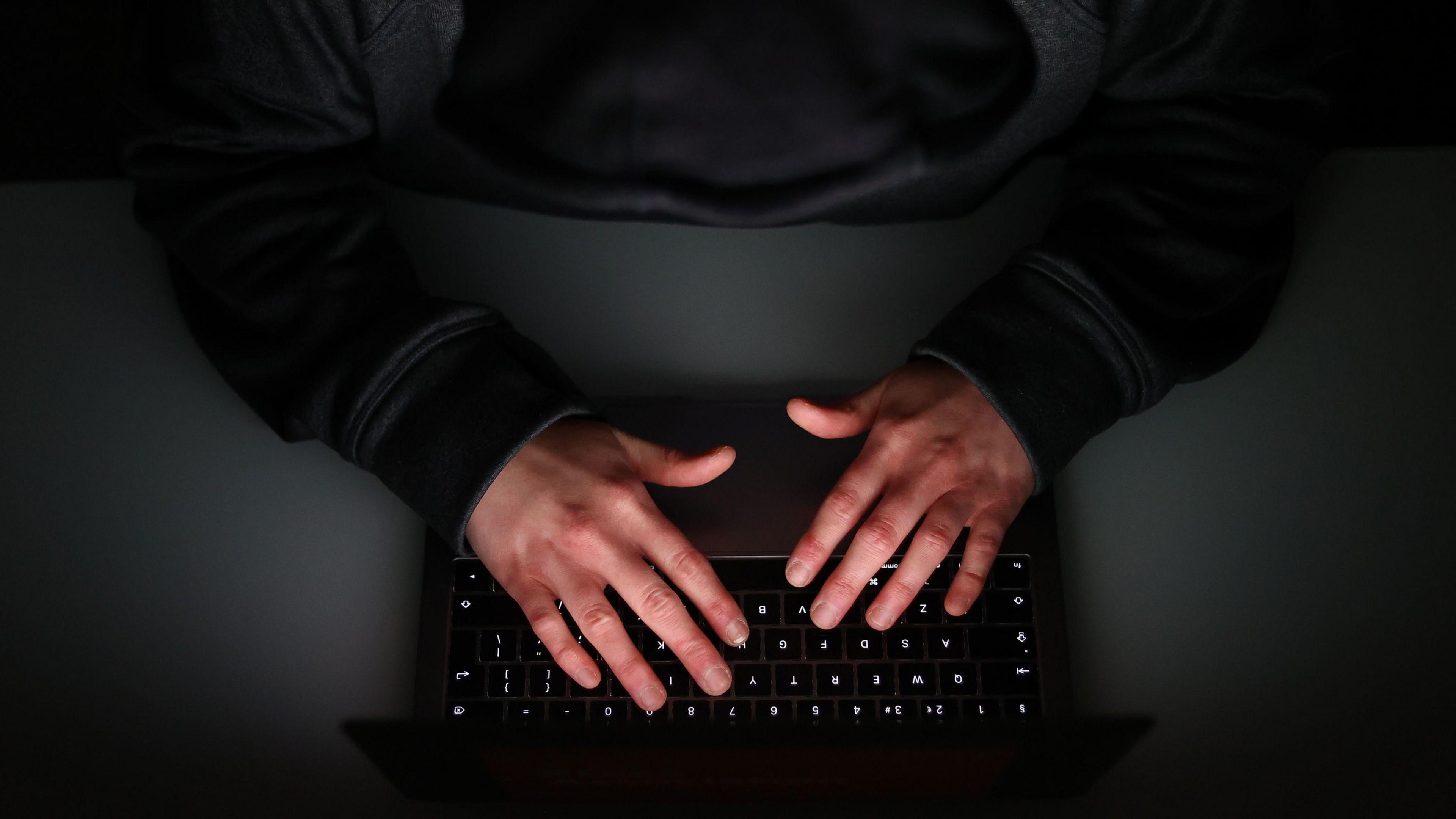 A pair of hands are positioned over a laptop keyboard on a desk. The identity of the person remains anonymous as only their arms are visible in the image.