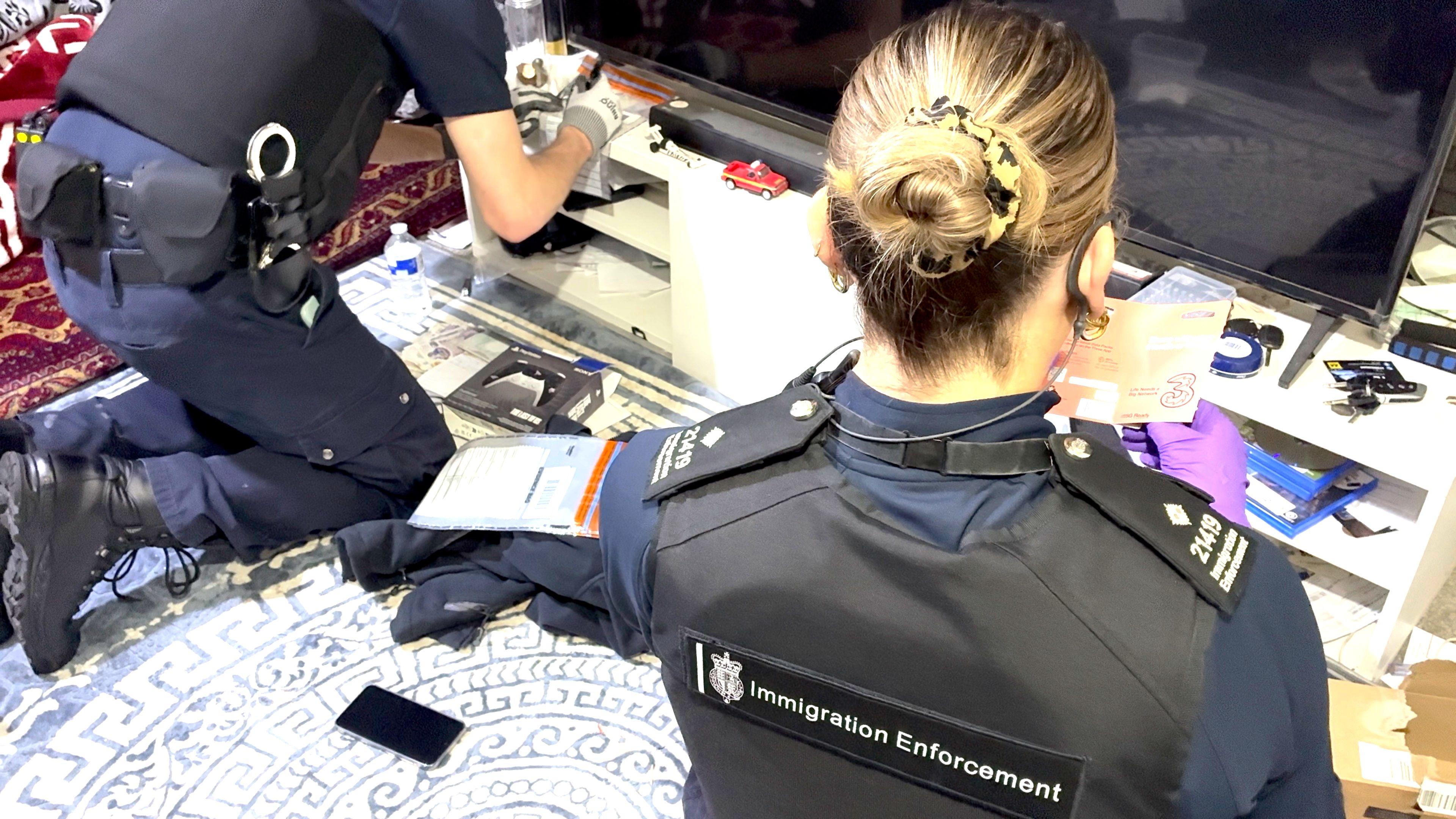 The back of two immigration enforcement officers look through belongings in a house