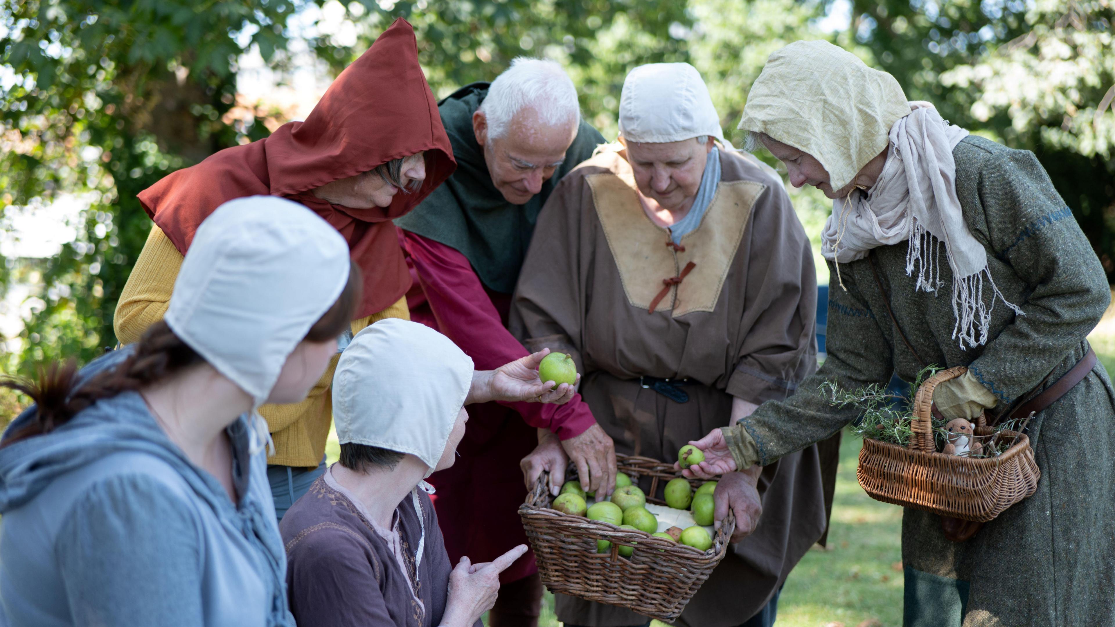 A group of actors dressed in medieval costumes, looking and pointing at baskets of fruit and herbs