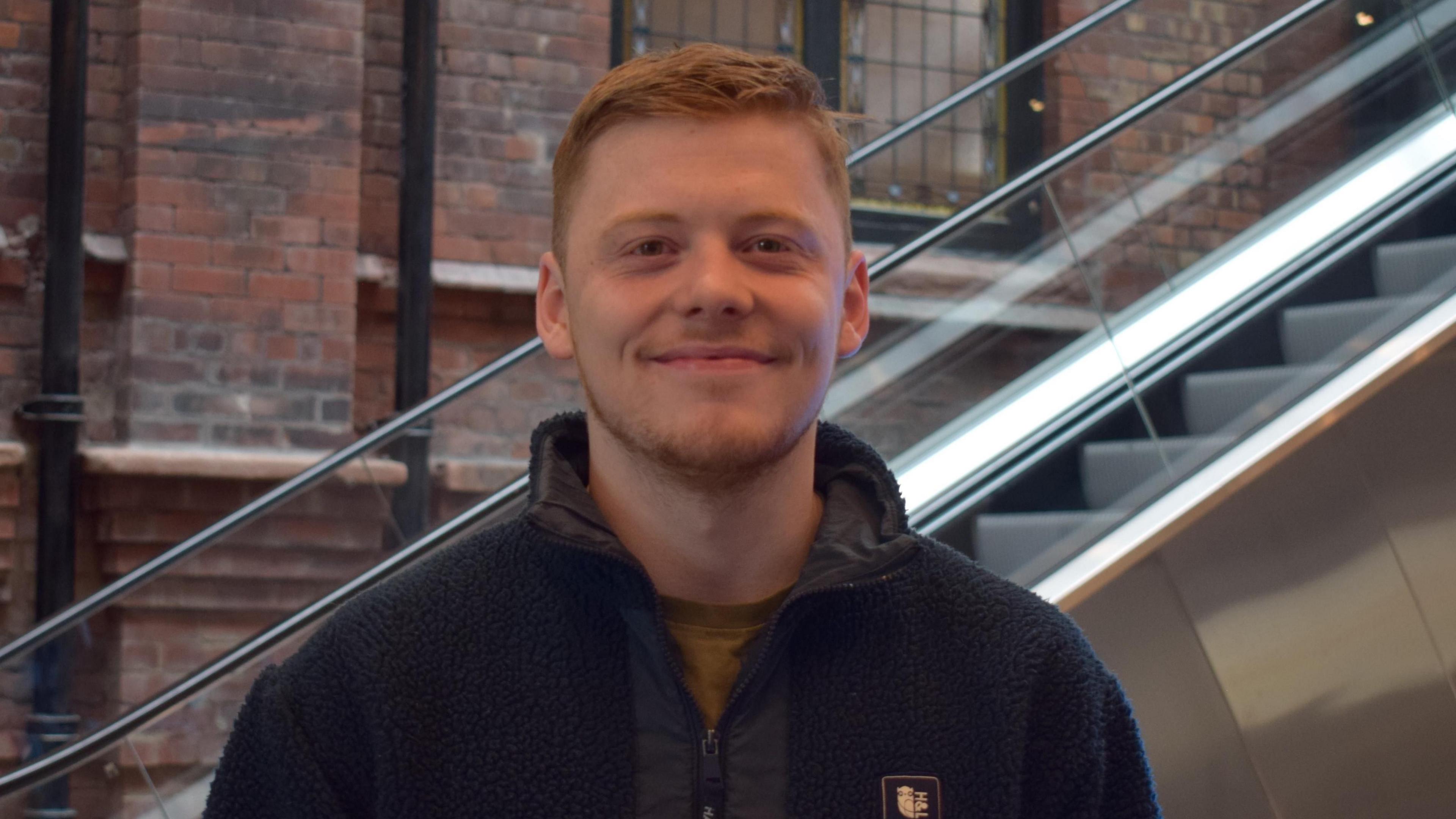 Ben stands smiling. He has ginger hair and a ginger beard. He has a blue fleece on. There is an escalator and a brick wall behind him.