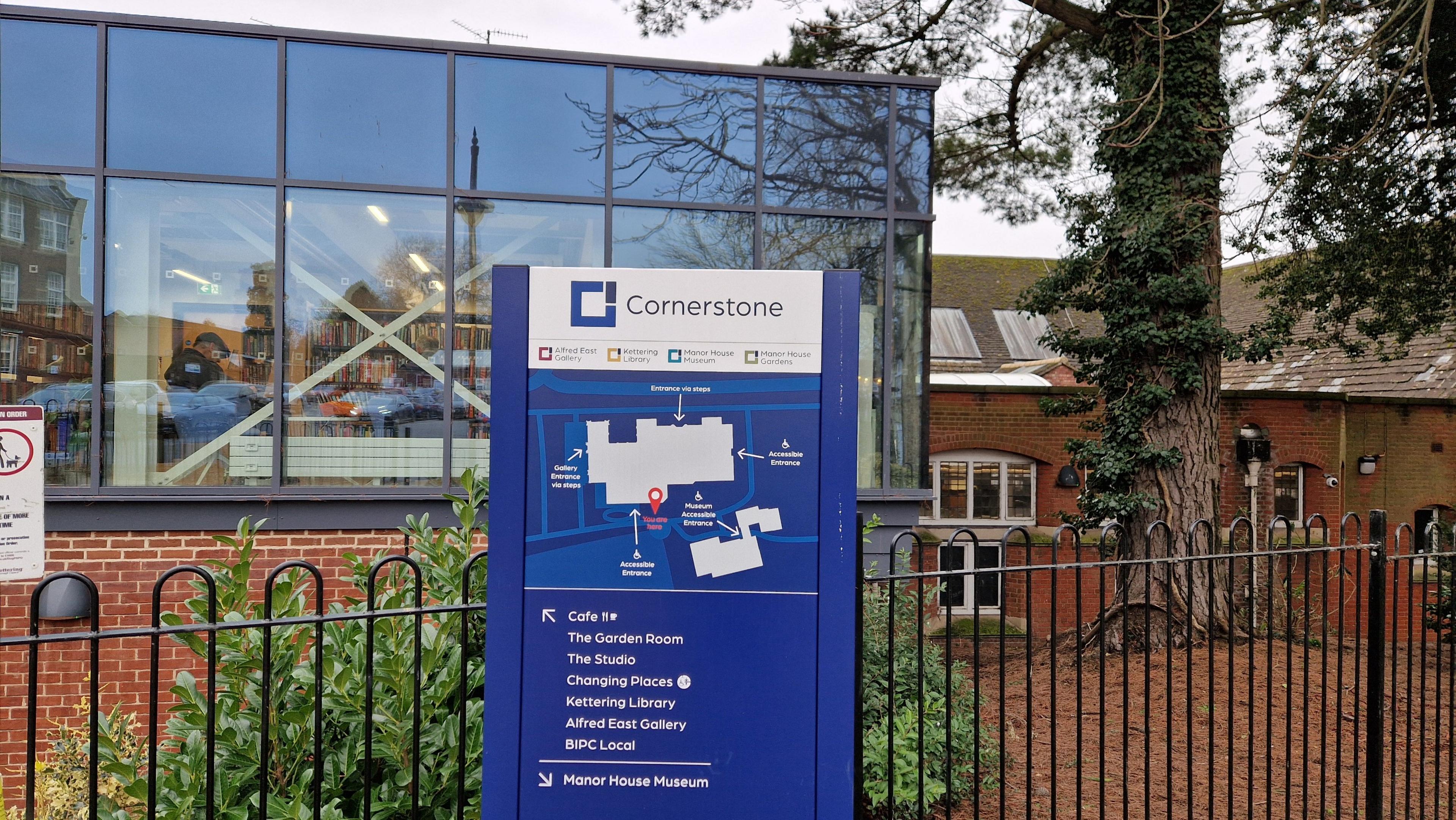 A single-storey red brick building with a large window revealing storage shelves within. A blue sign outside says "Cornerstone" and shows a map of the site. There are railings behind the sign.