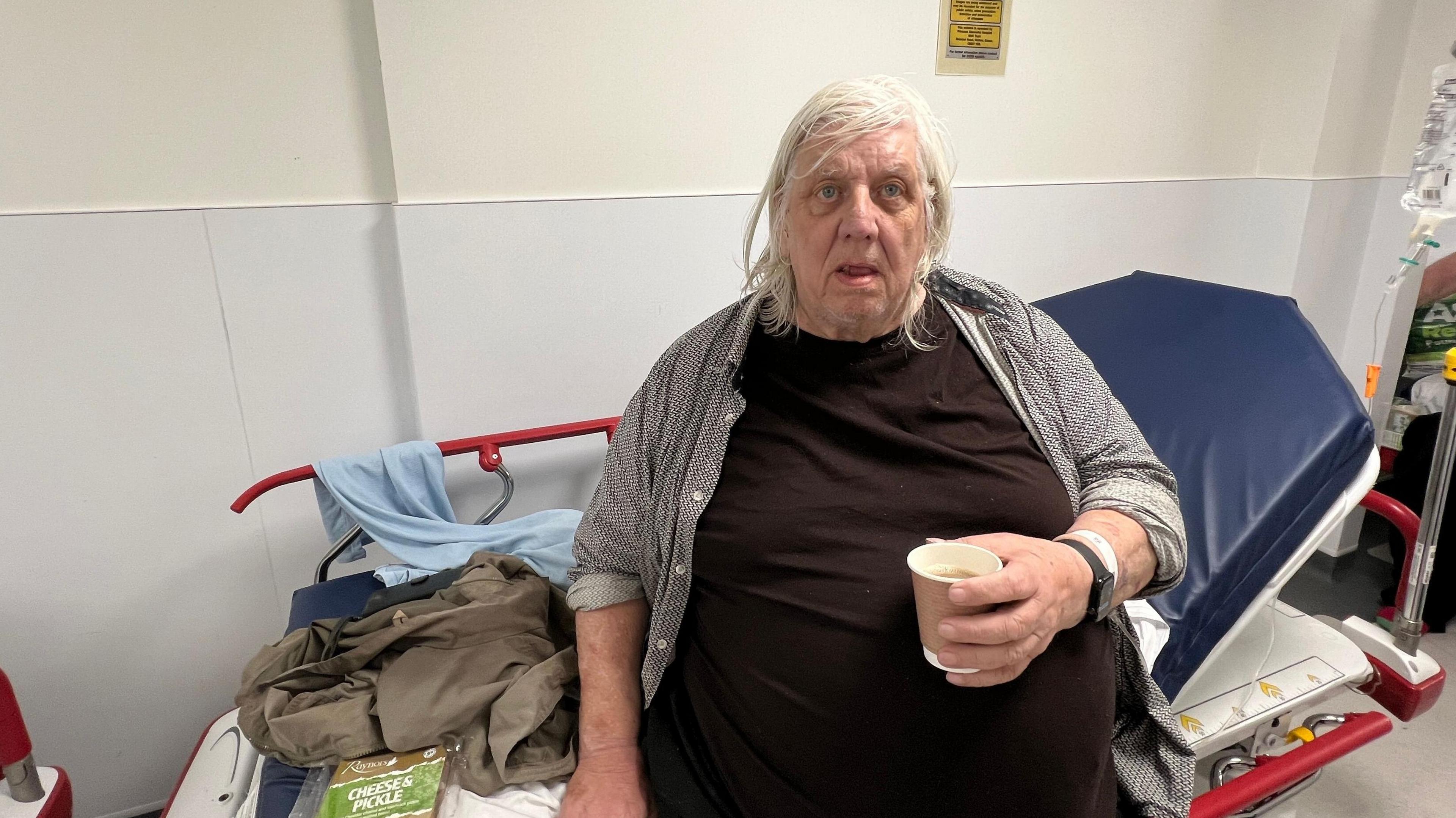 Patient waiting on a hospital trolley in A&E with a paper cup of tea in his hand.