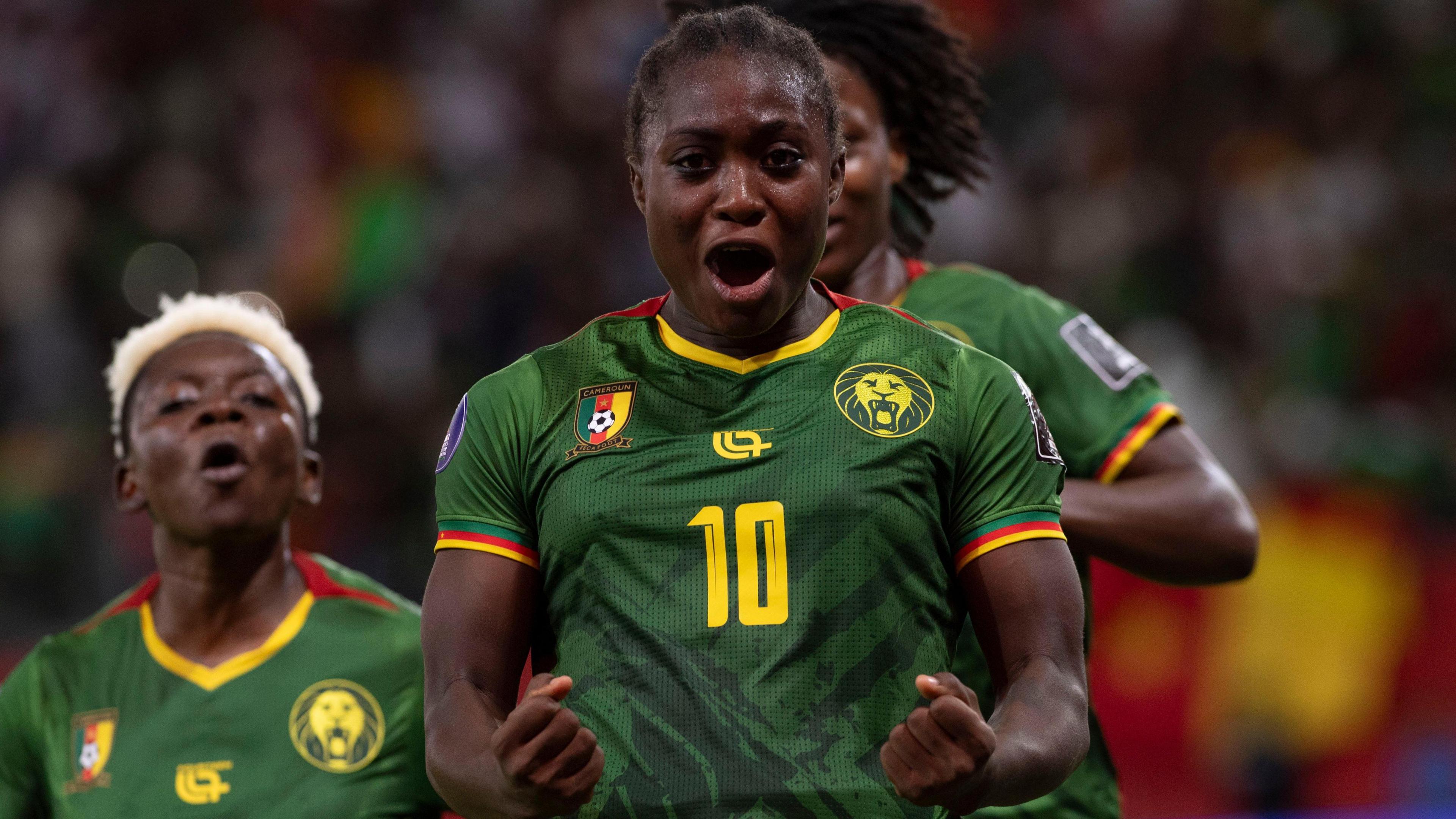 Naomi Eto, wearing a green Cameroon shirt with a yellow number 10 on the chest, clenches her fists down by her waist and shots into the camera in celebration