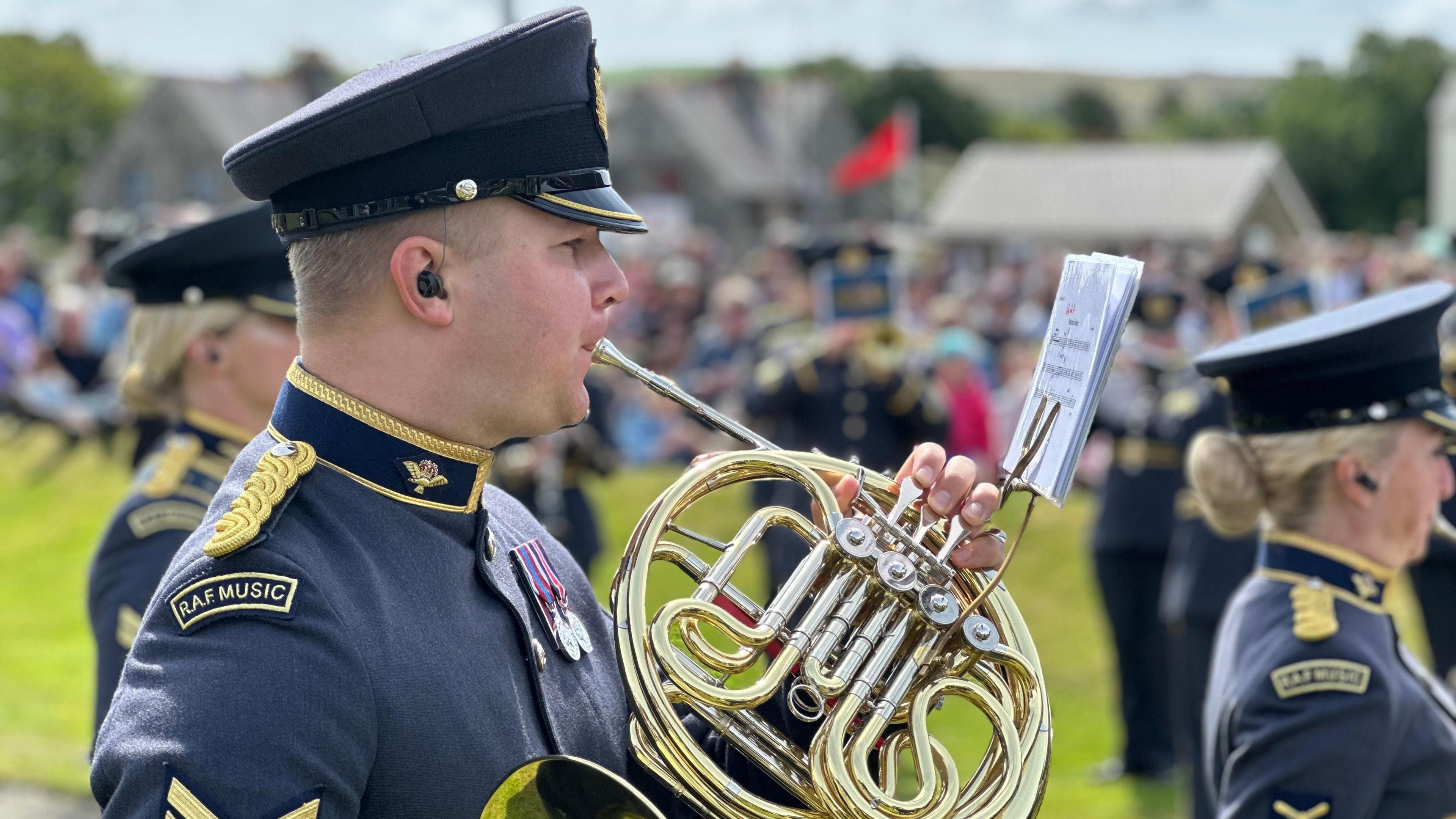 A members of the Band of the RAF Regiment plays a shiny brass instrument while reading music off a stand attached to it. His uniforms is grey-blue with gold trim and RAF Music is stitched into a badge on his shoulder.