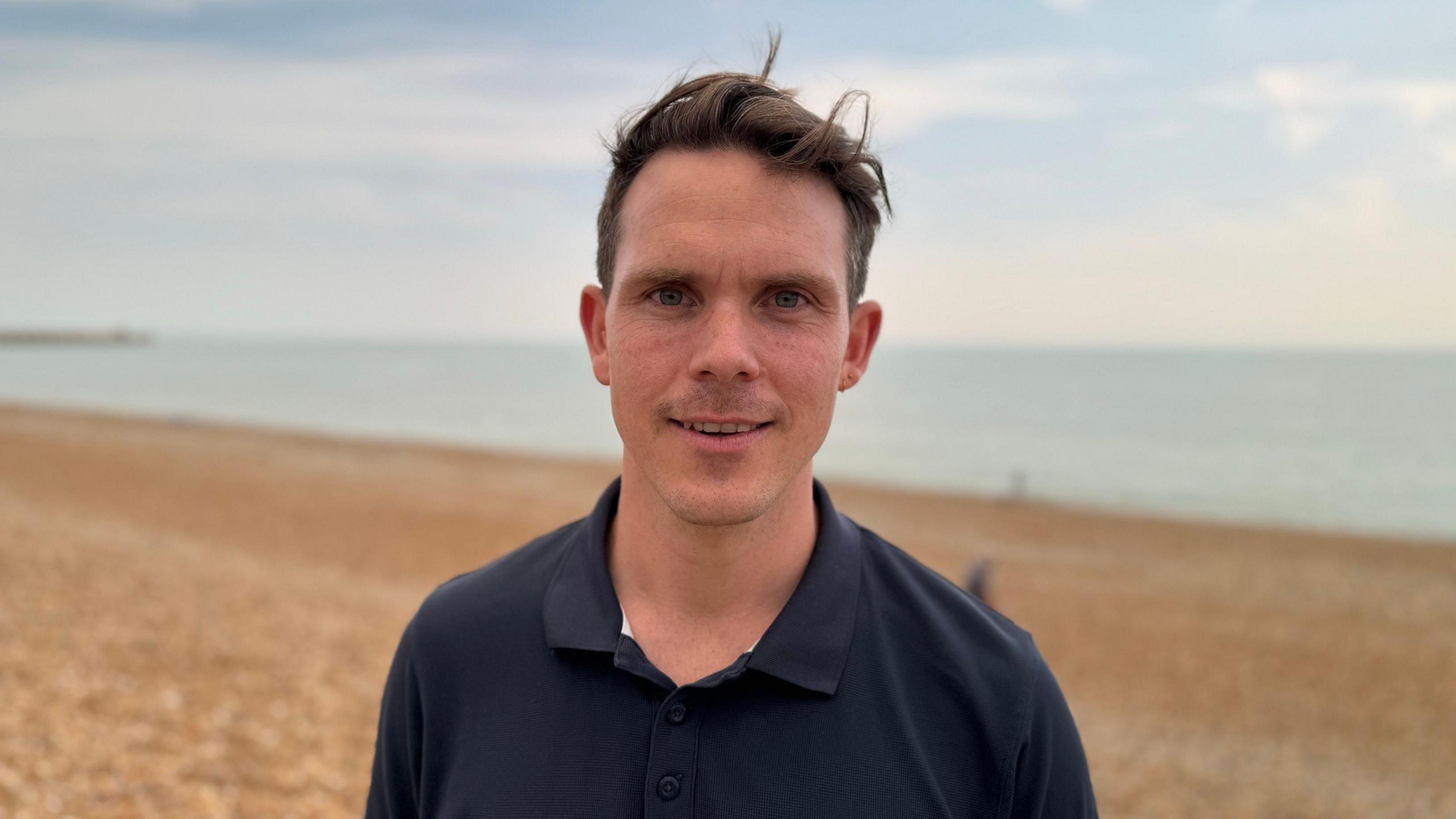 Sam Johnson who has brown short hair and a black polo top. Sam is standing on Brighton beach which has a shingle bank
