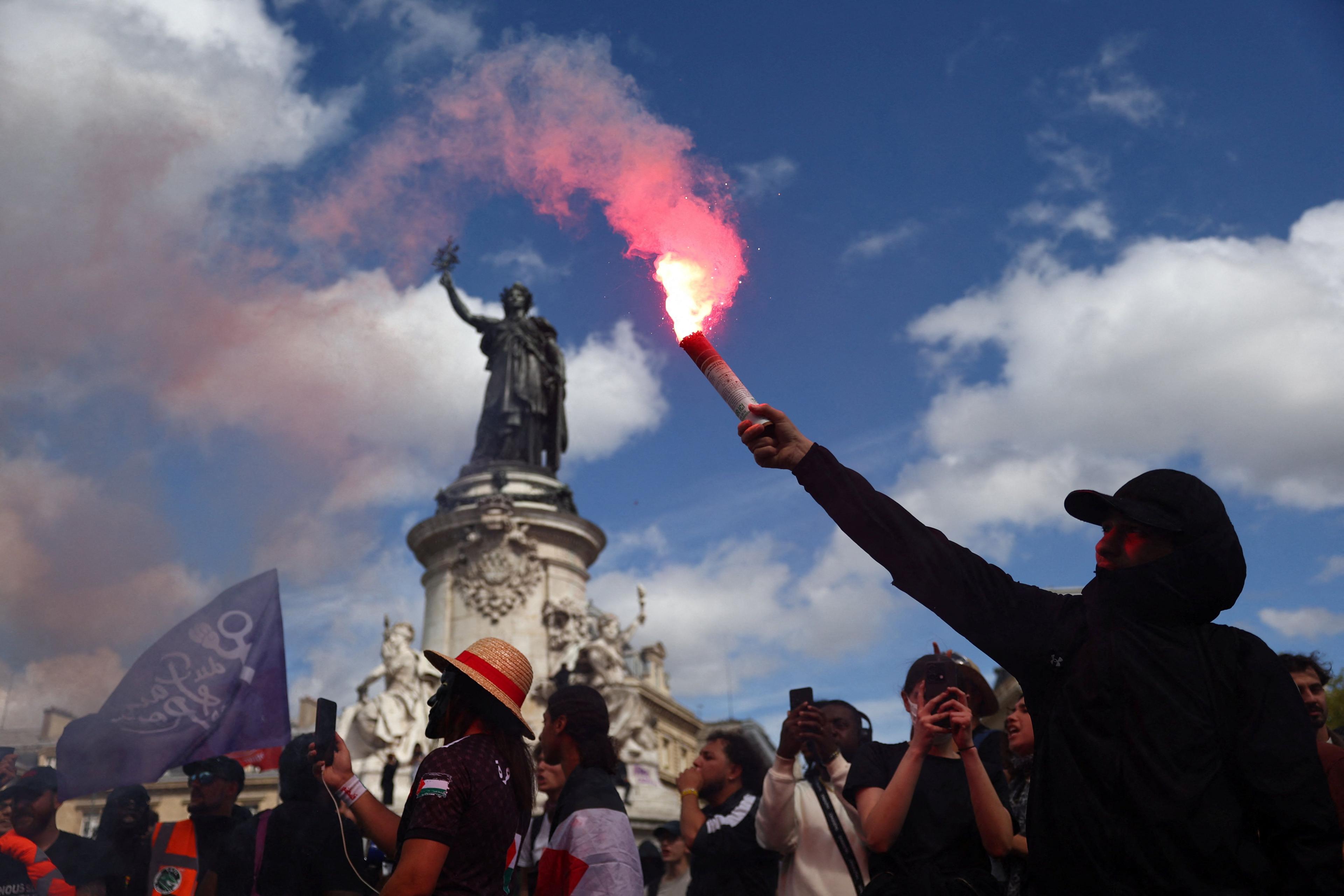 A man holds a flare aloft against the backdrop of a statue, a blue sky and crowd