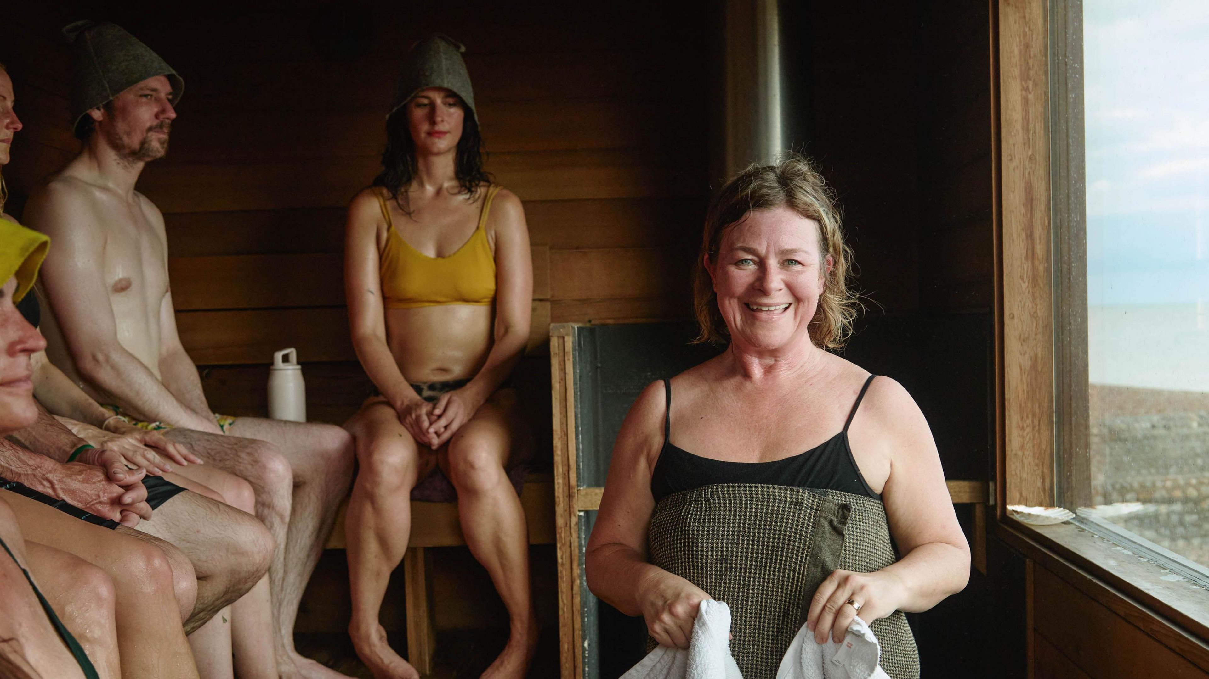 Liz Watson looks at the camera holding a towel and wearing a black swimming costume. She stands inside a wooden sauna with four other people in shot, who are all wearing swimming costumes and hats. There is a window looking out on the sea. 