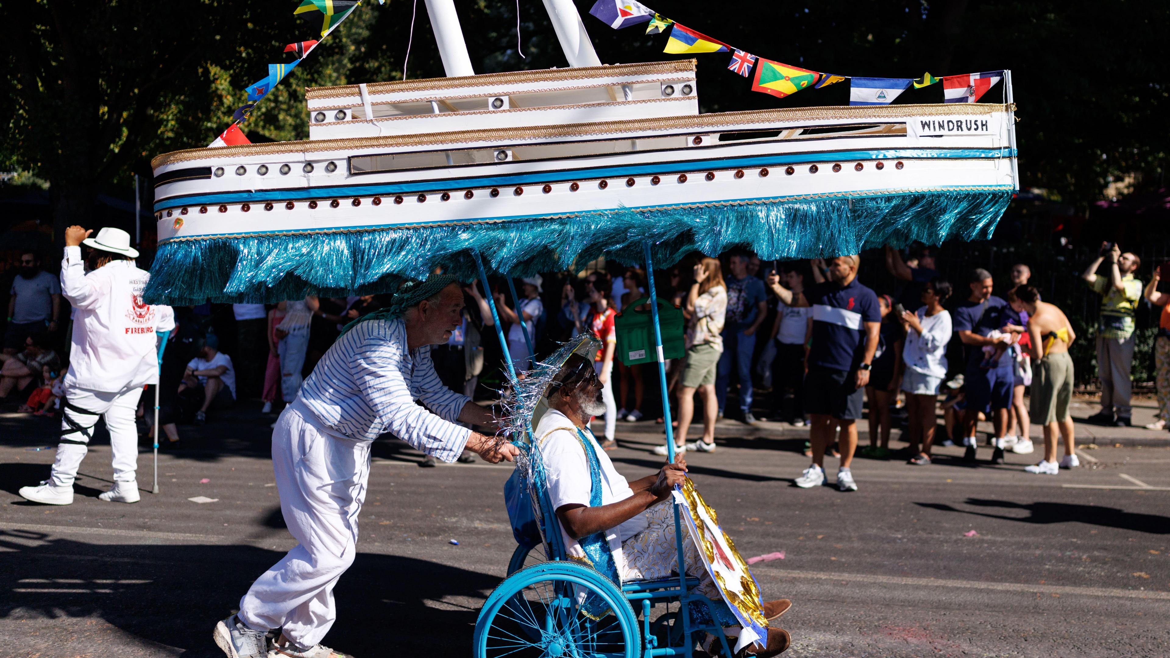 A man wearing white is in a blue wheelchair. Attached to the chair, above him, is a large model boat labelled: Windrush