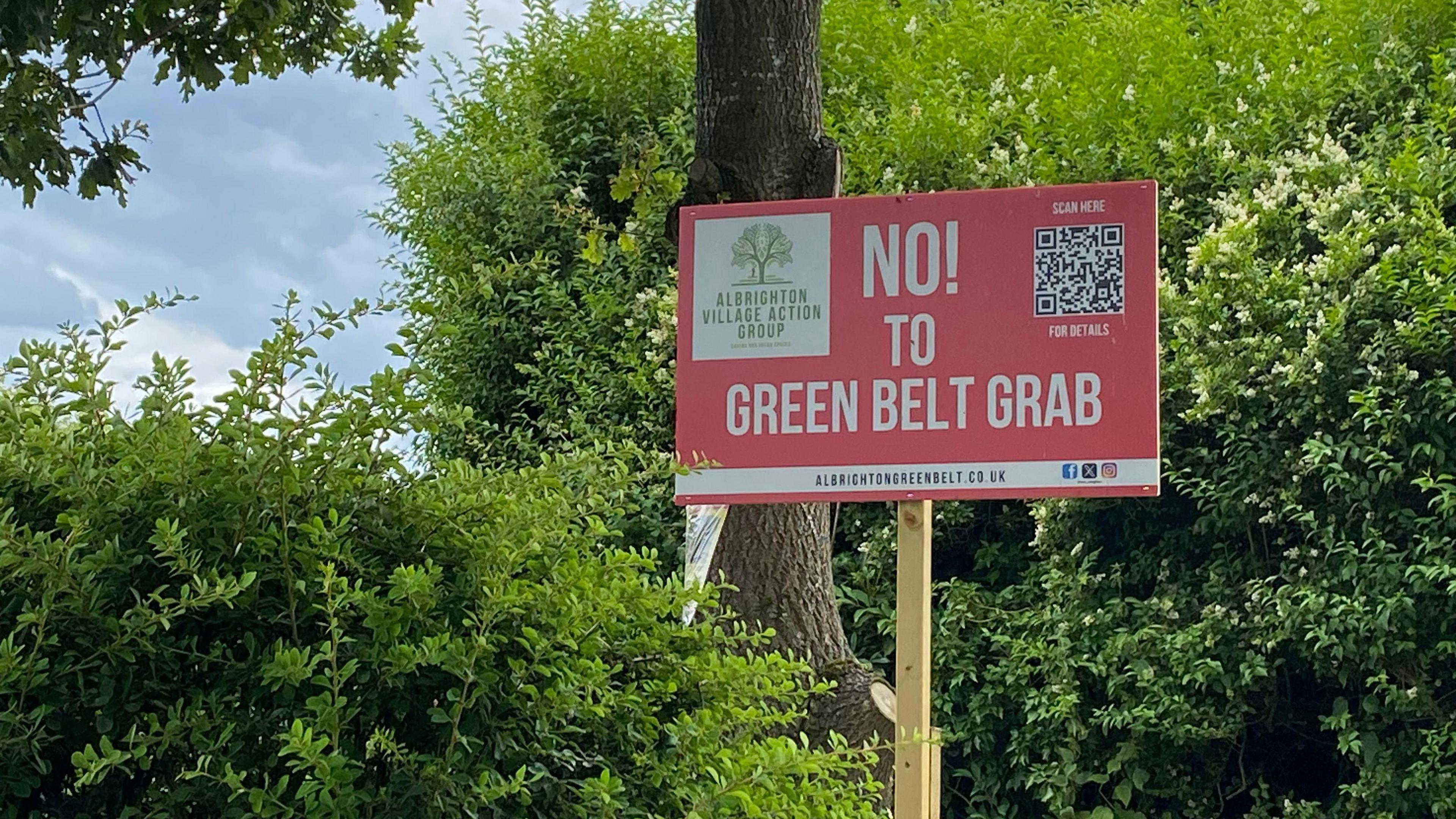 A red placard with a wooden stake in a rural location, with the words "No to green belt grab" written in white writing