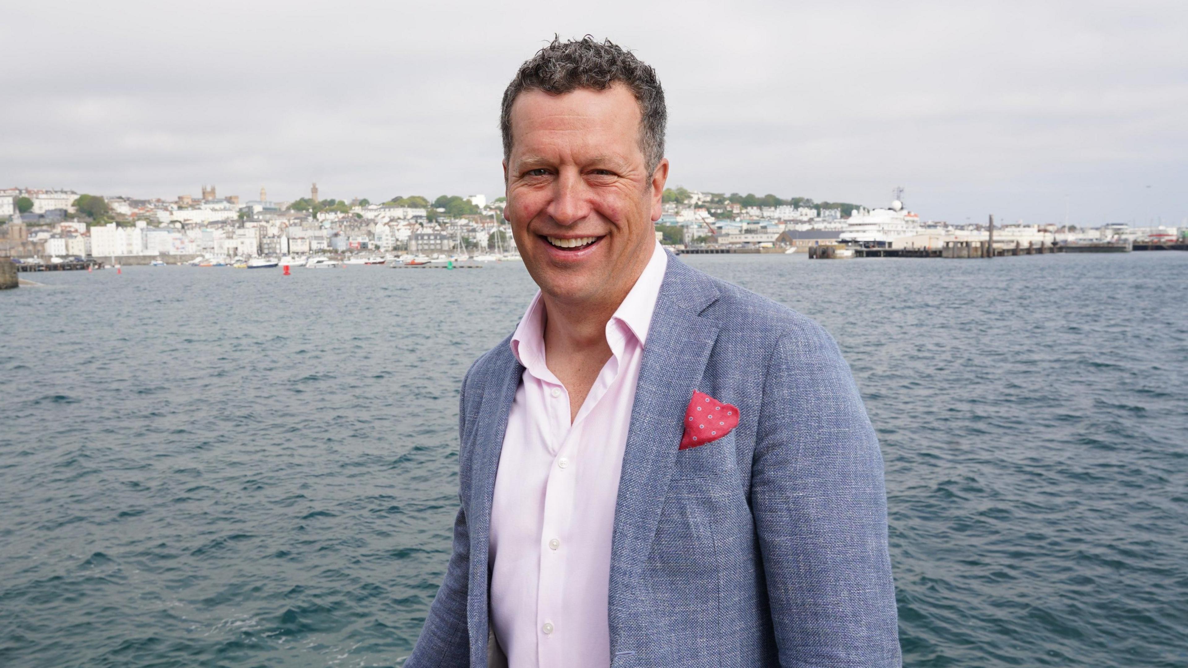 Jay Goss is pictured smiling at the camera, against the backdrop of the sea. He is wearing a smart, grey suit jacket, with pale pink shirt underneath. He has brown hair and is pictured amidst the backdrop of the sea, with a boat and housing in the background.