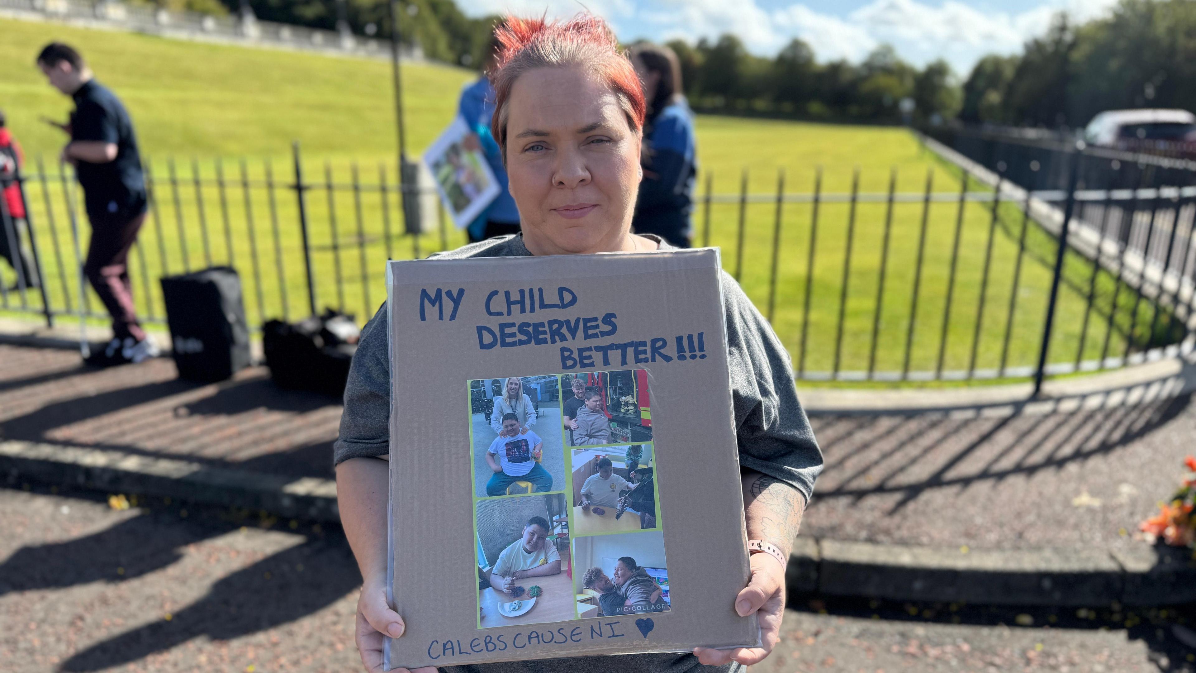 Tina Henderson has red hair tied in a bun holds a cardboard sign which reads "My child deserves better." She wears a grey t-shirt, behind her is a black metal fence with a patch of green behind her.