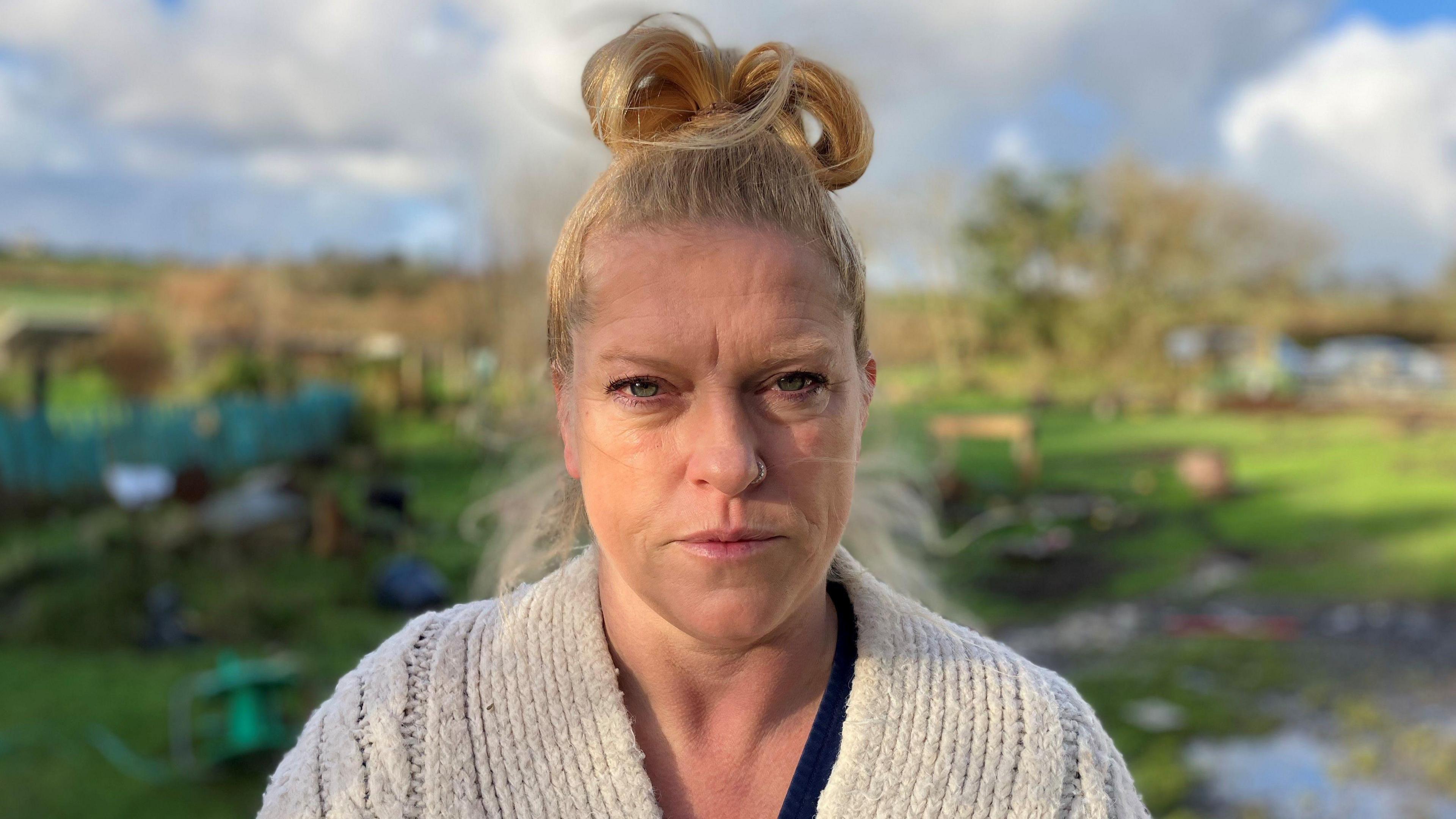 Woman with her hair tied up in a pony-tail on top of head and wearing a cream pullover with garden behind 