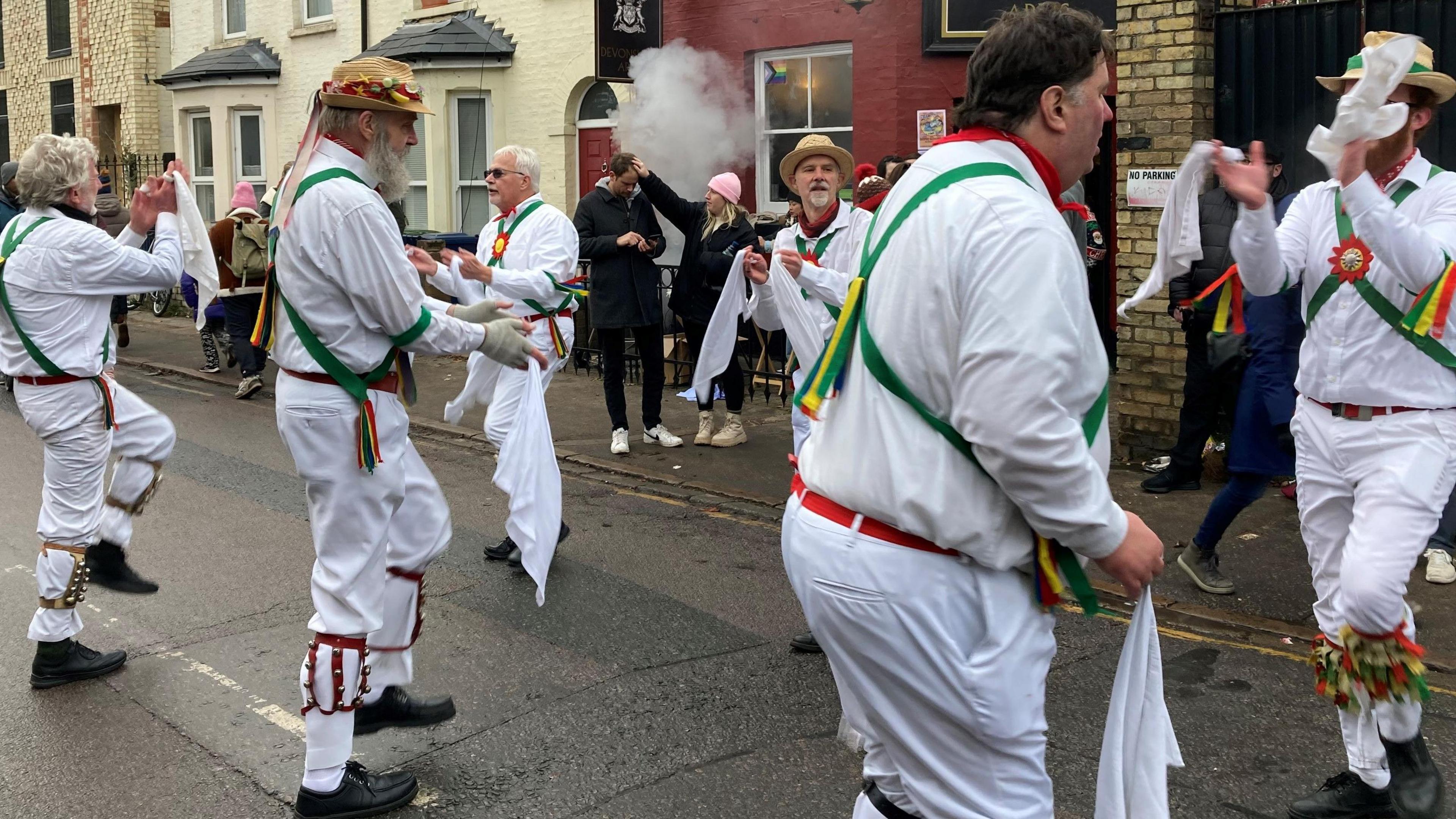 Morris Dancers on Mill Road in Cambridge. They are wearing traditional white trouser and shirts. They have bells on their legs and are waving white handkerchiefs around. There are people walking by in the background