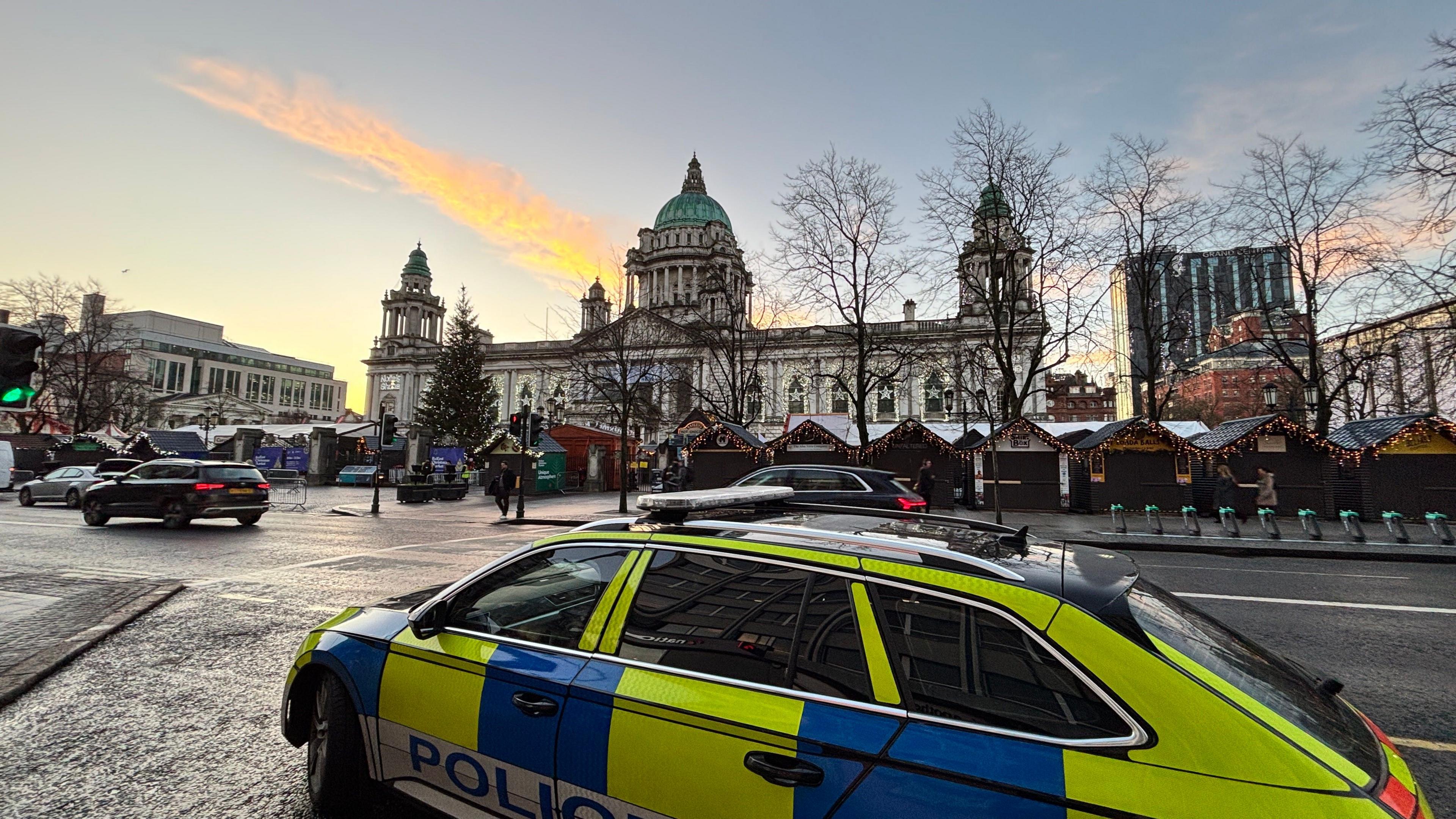 Police car in front of Belfast City Hall. There are cars and market stalls in front of the city hall. The sky is blue.