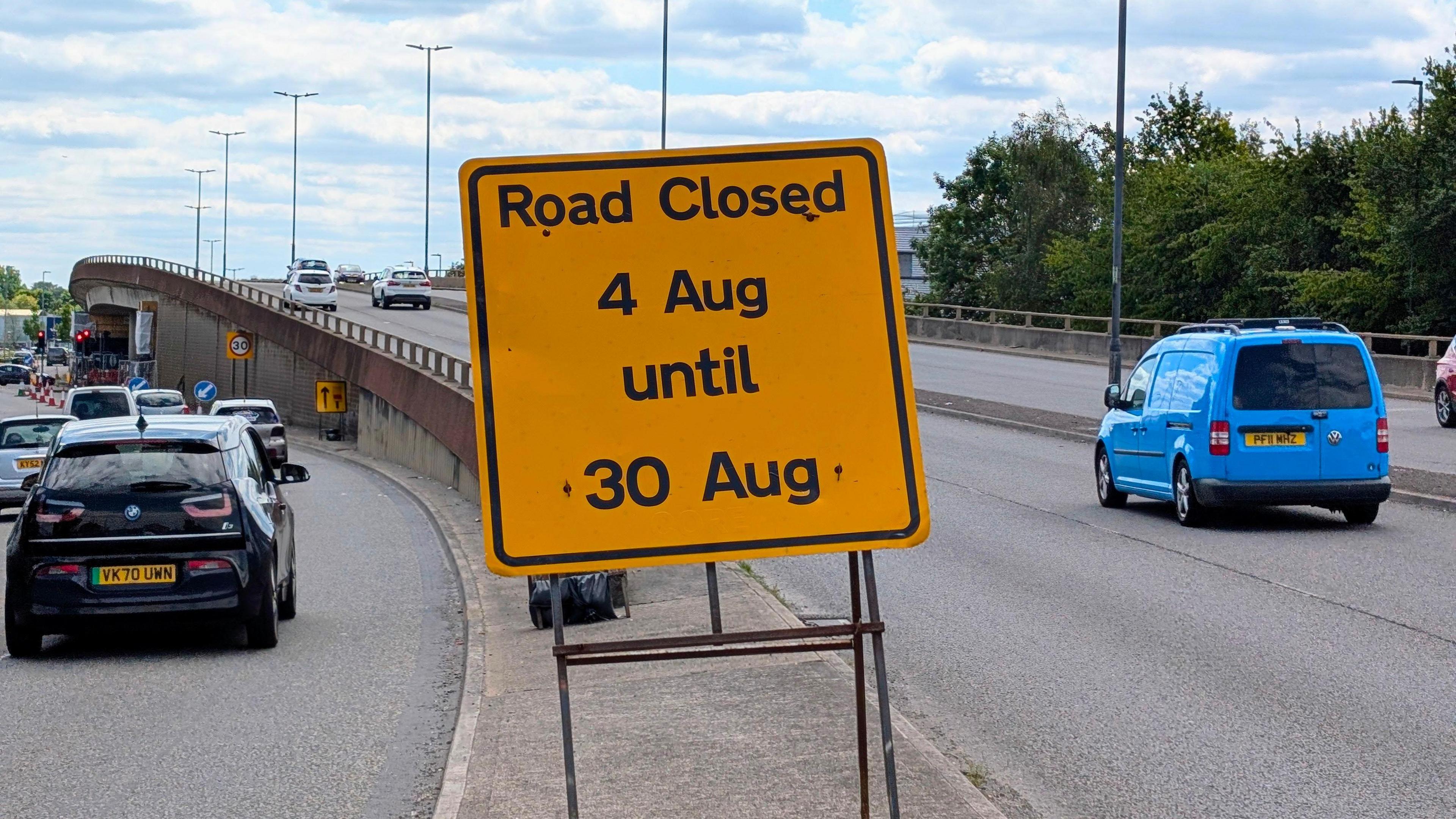 A view of the flyover with cars driving over it. Several drive to the side of it. A yellow sign in the middle says "Road closed - 4 Aug until 30 Aug".