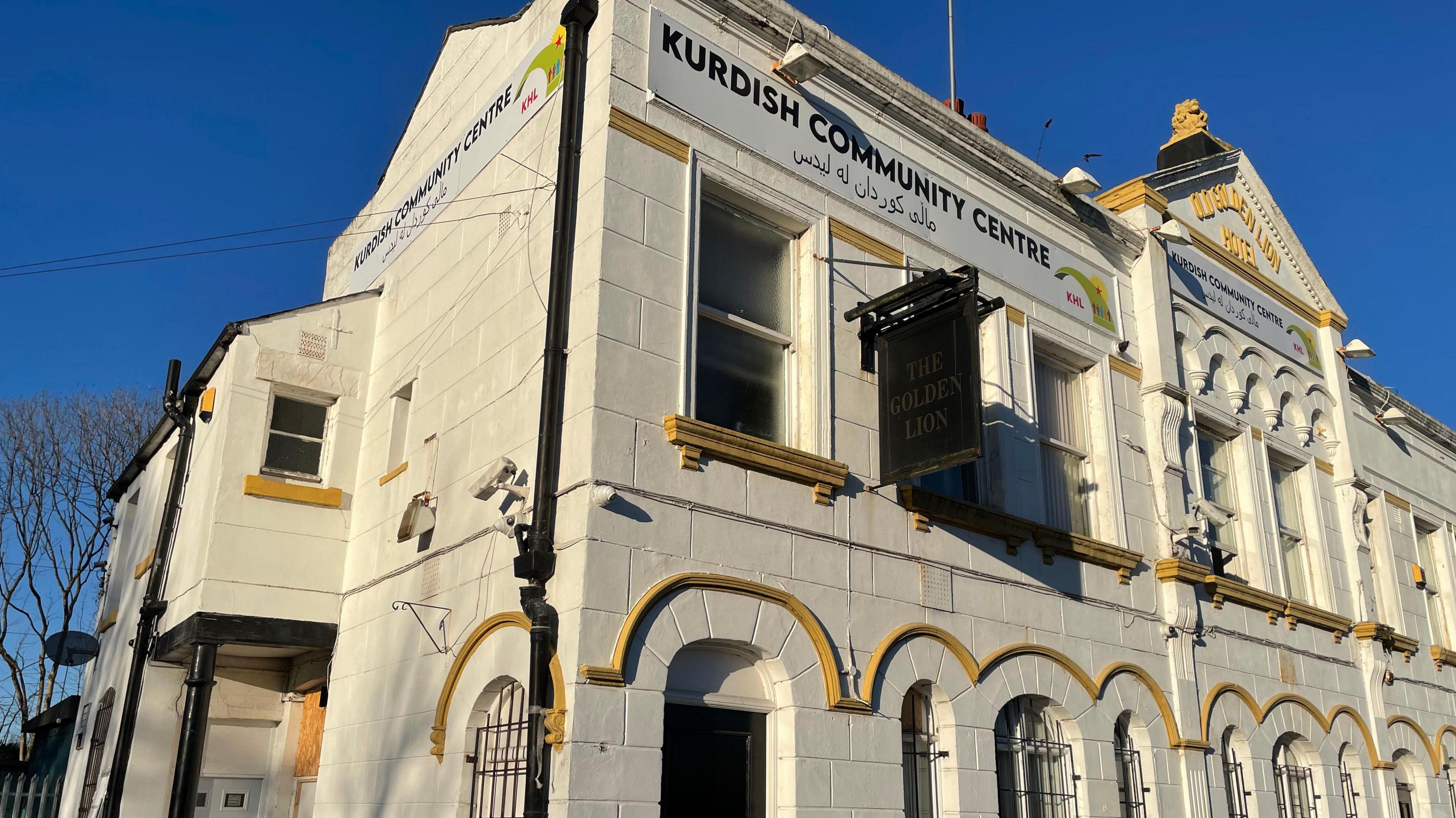 A white building with yellow lines around the window frames stands in the sunshine. It is labelled 'Kurdish Community Centre' above a black sign saying 'The Golden Lion'.