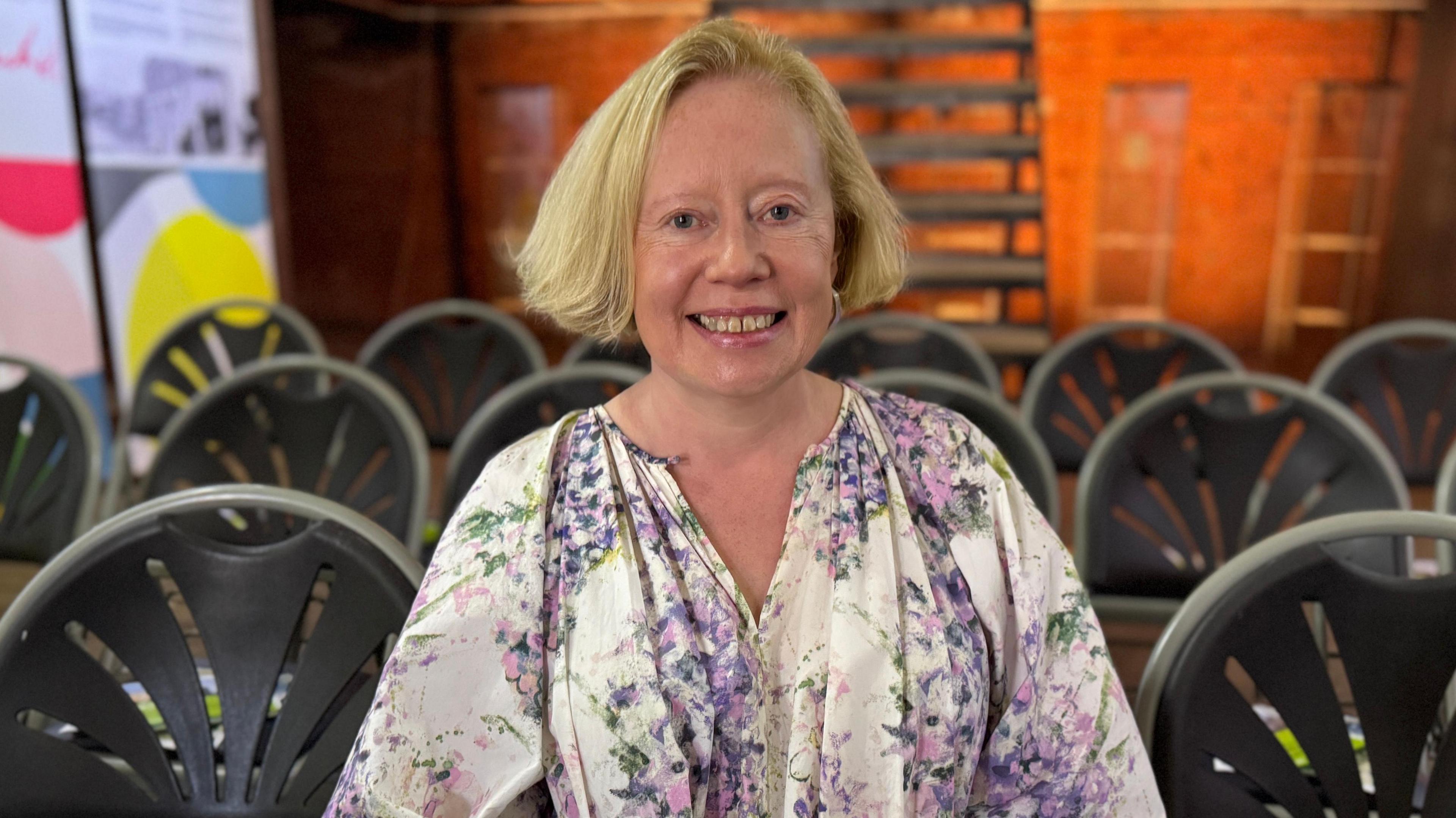 A woman smiles broadly at the camera. She has short blond hair and is wearing a purple and white top with a floral pattern. In the background we can see empty chairs set up for an event. 