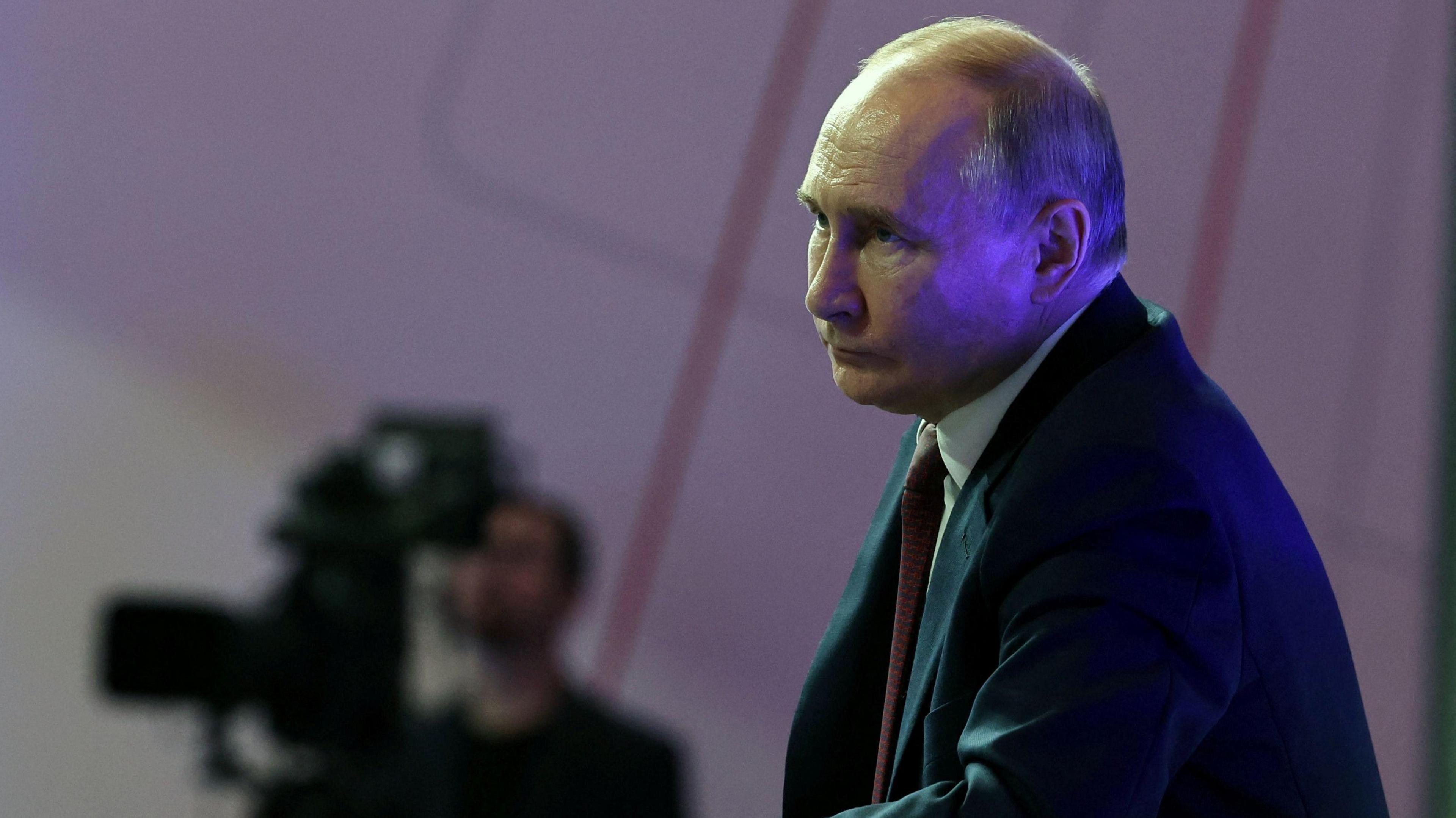 A side profile of Vladimir Putin with a stern expression, his face partially shaded