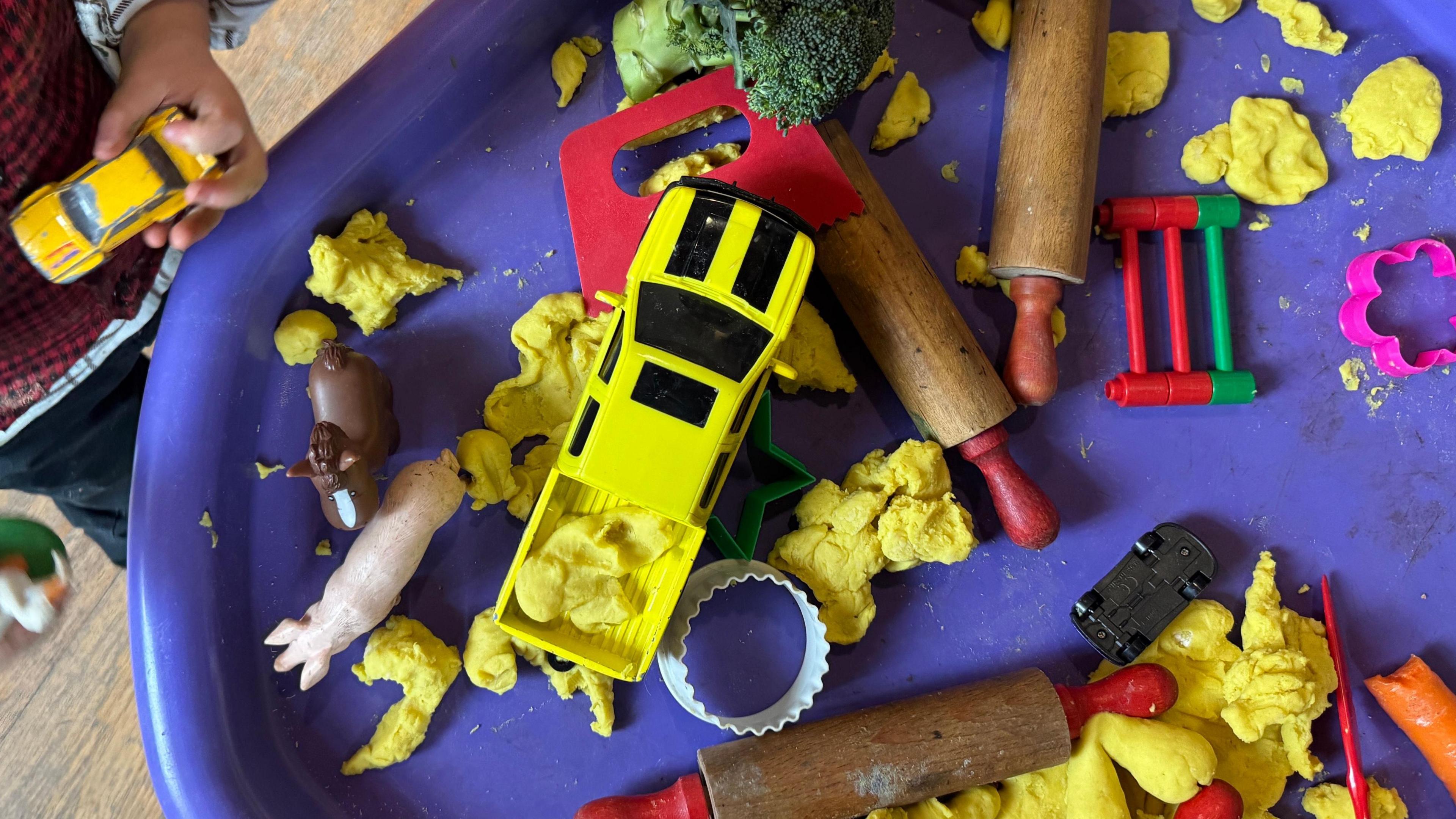 Clai gyda anifeiliaid a siapaiau wedi eu gosod ar fwrdd porffor gyda dwylo plentyn yn ymestyn am y tegannau. 