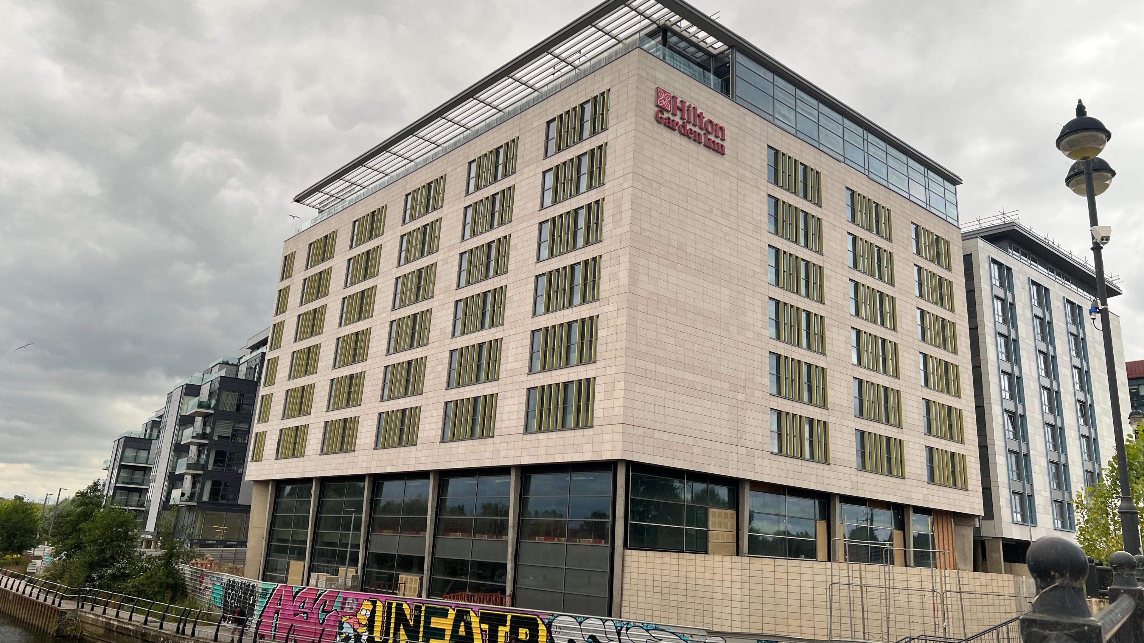 A tall beige building with a large glass ground floor and 6 storeys high. Teh words 'Hilton Garden Inn' are at the top and a graffiti covered fence is in front, with a river in front of that. 