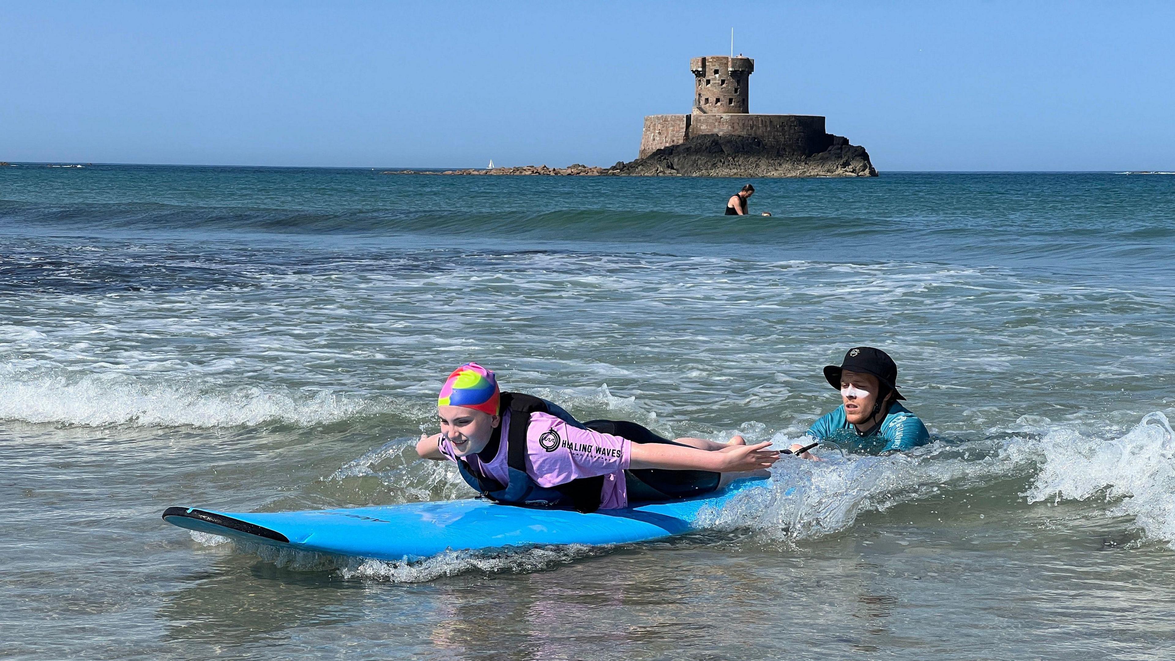 A girl body boards on a small wave while a volunteer helps push her long and there's a castle in the background