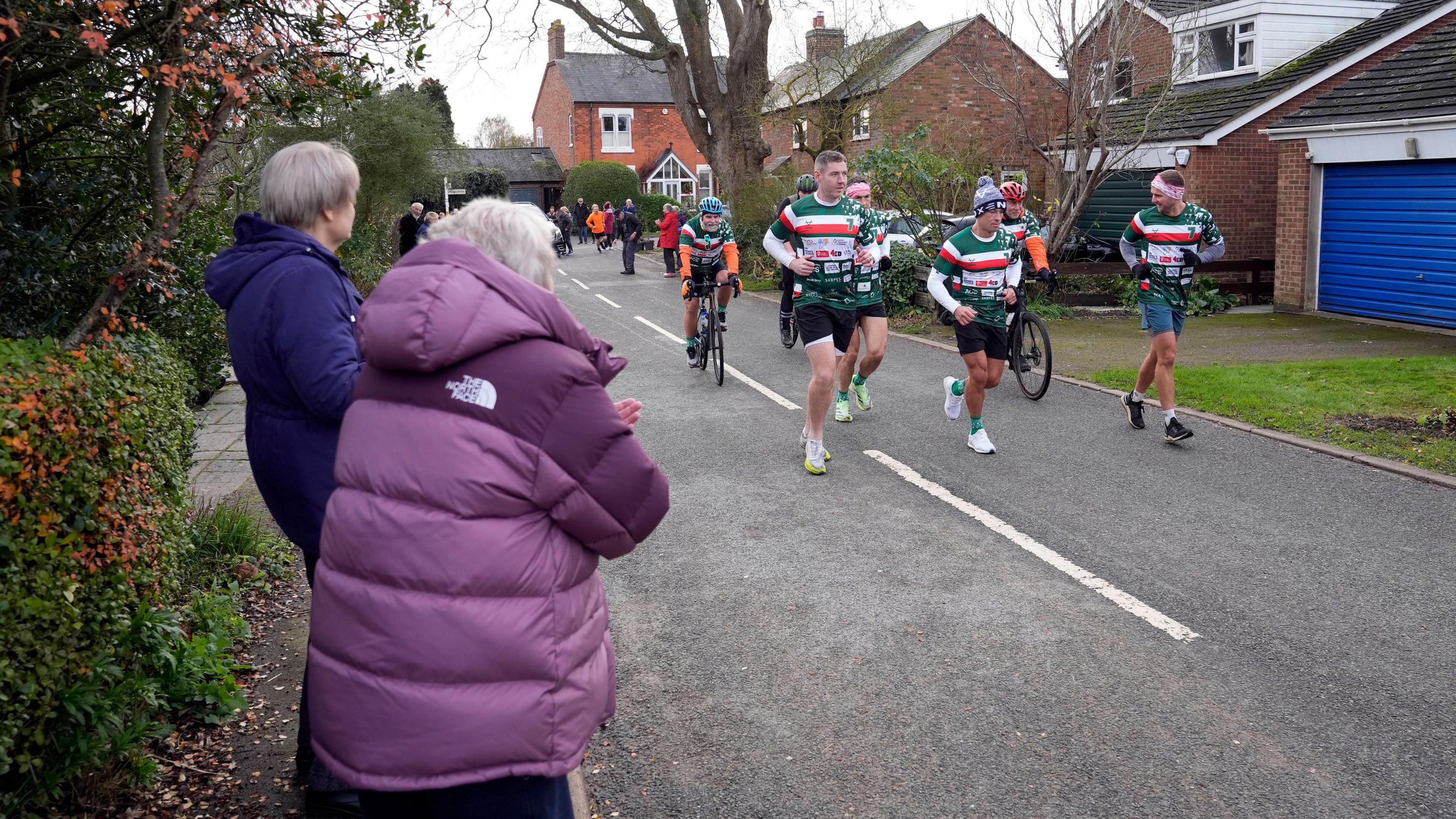 A man in a blue jacket and a woman in a purple jacket clap as a pack of runners wearing green, red, white striped rugby shirts run down a road of a housing estate.