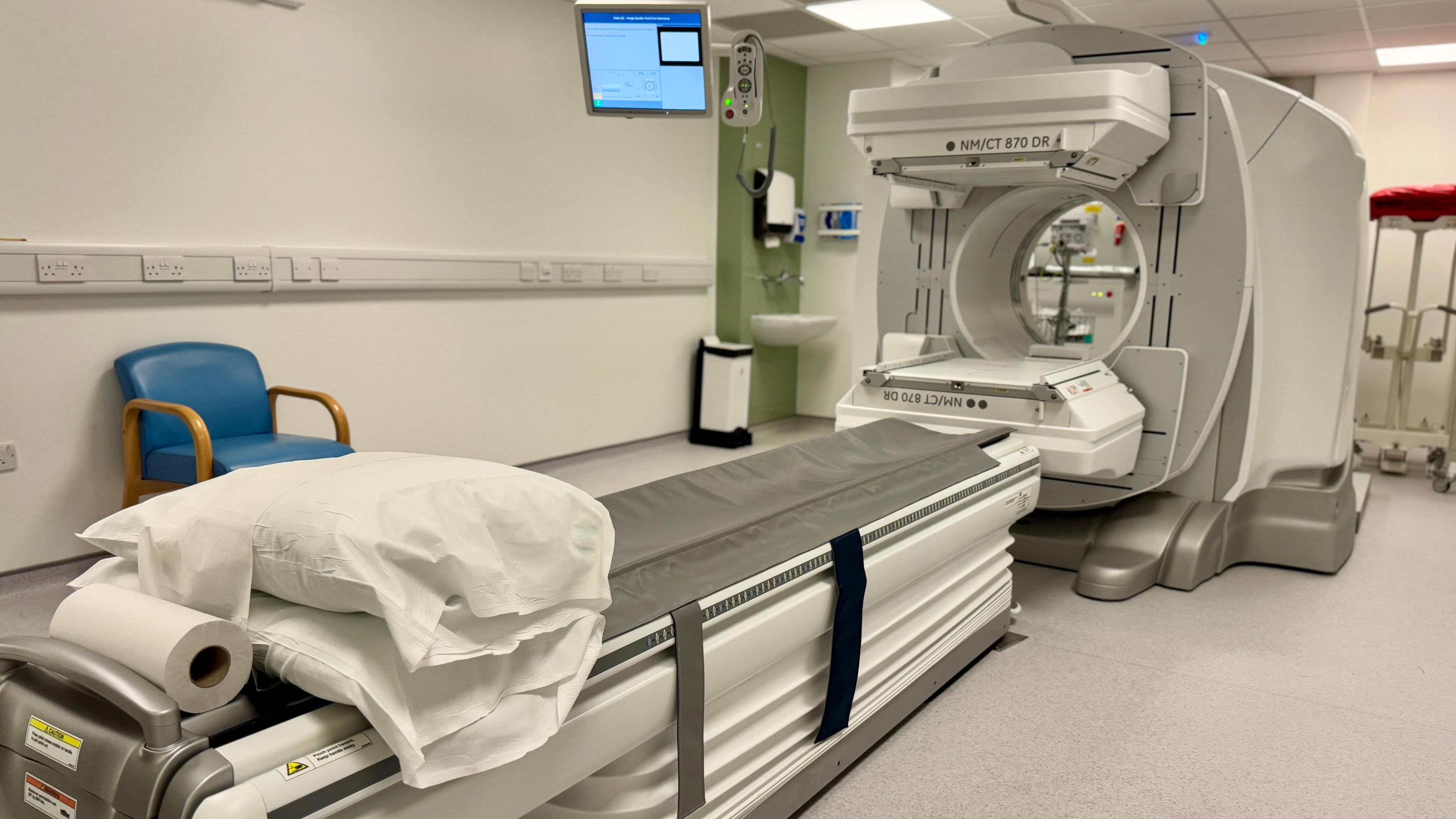 View of the new combined gamma and CT scanner. A flat bed with two white pillows on it is at the front of the shot with a circular scanner at the foot of the bed. 