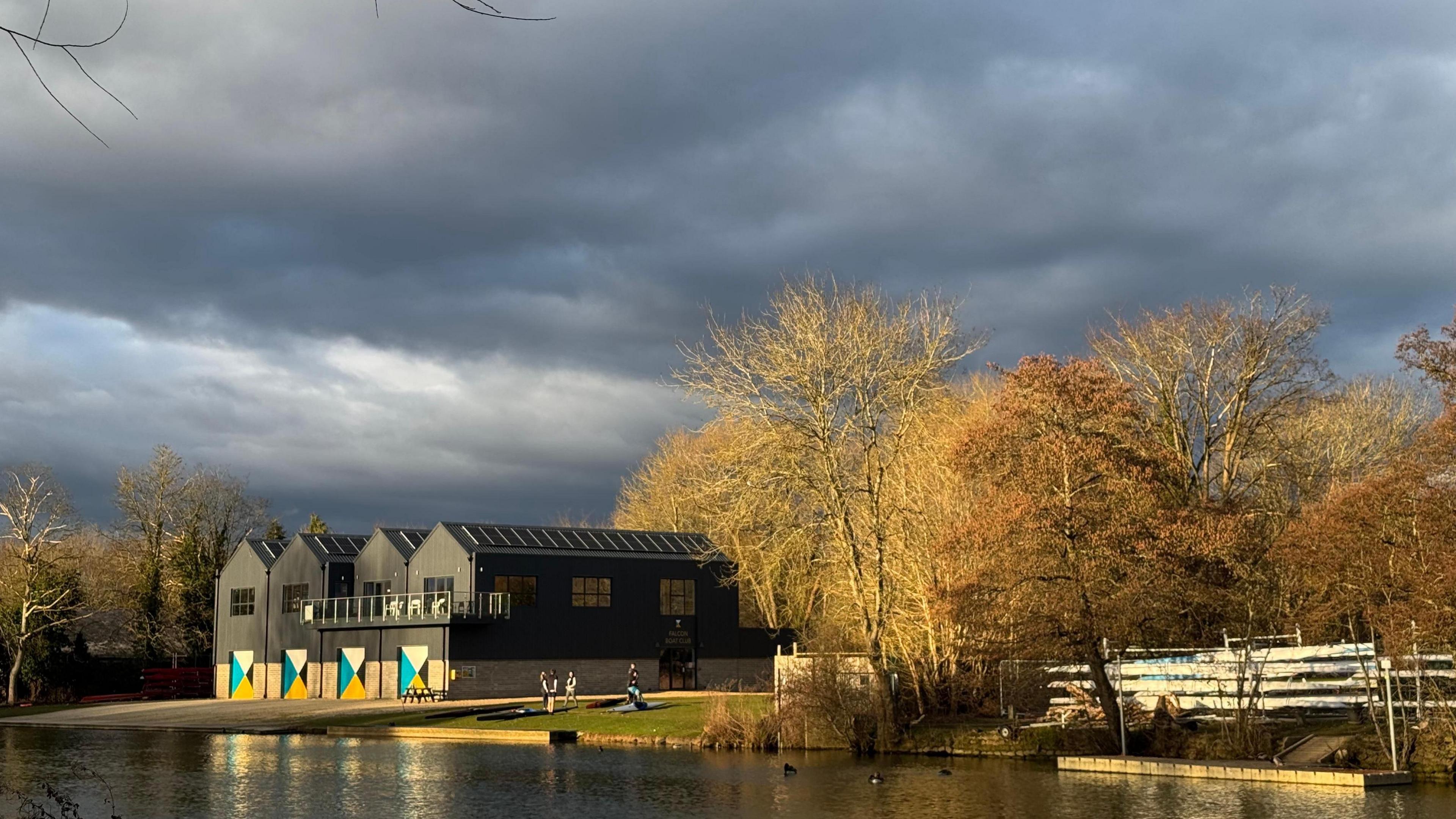 A large black boat house with colourful doors stands on the bank of the River Thames. The building is in sunshine, but overheard there are dark black clouds. On the banks of the river are leaf-less trees.