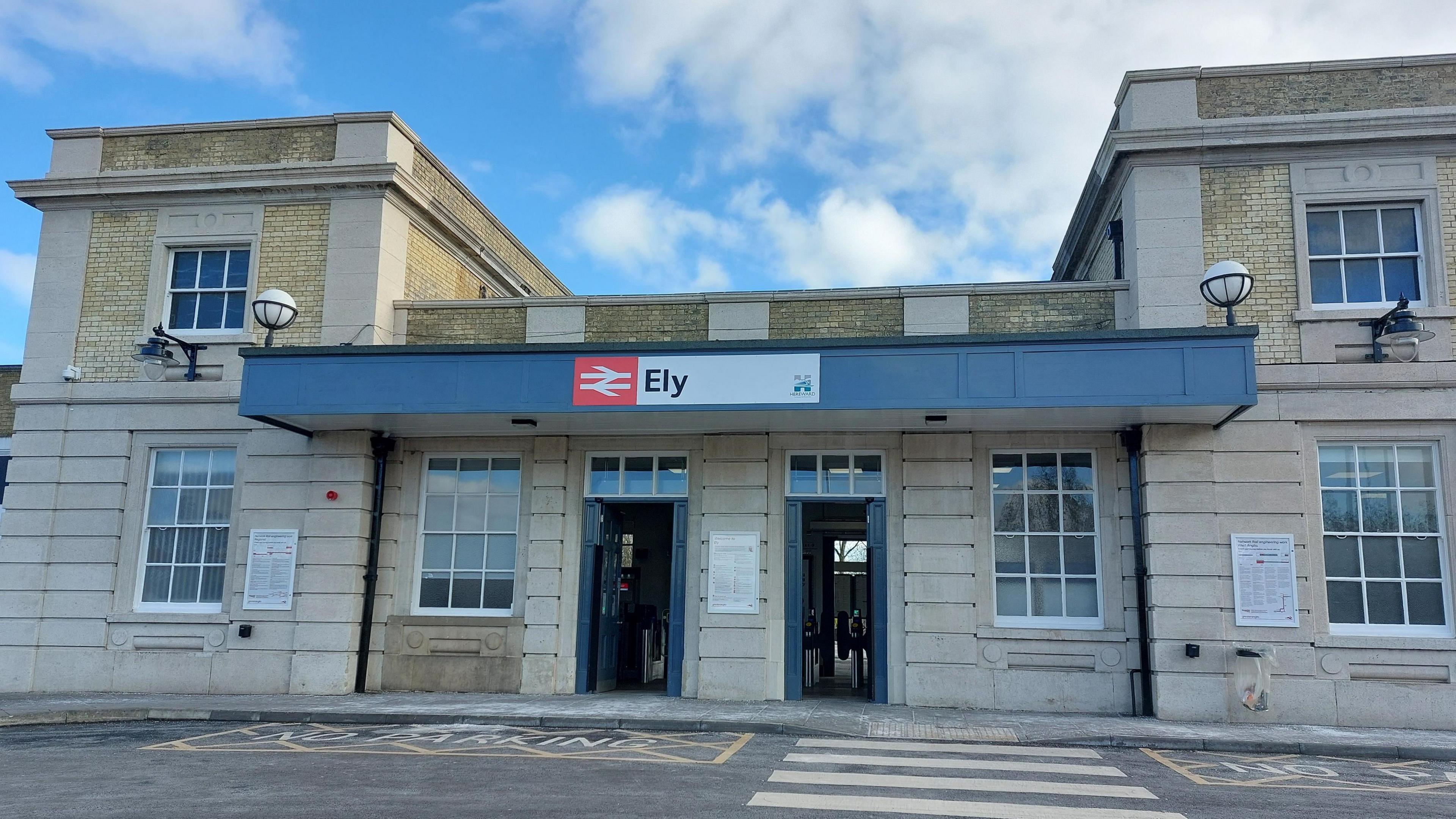 The exterior of Ely railway station. It is an early Victorian building, made of yellow brick and stone. There are square two-storey wings on either side of the single-storey entry, which has two doors and two large sash windows. Above the doors and windows is a grey painted sign saying Ely with the National Rail symbol, white on red. 