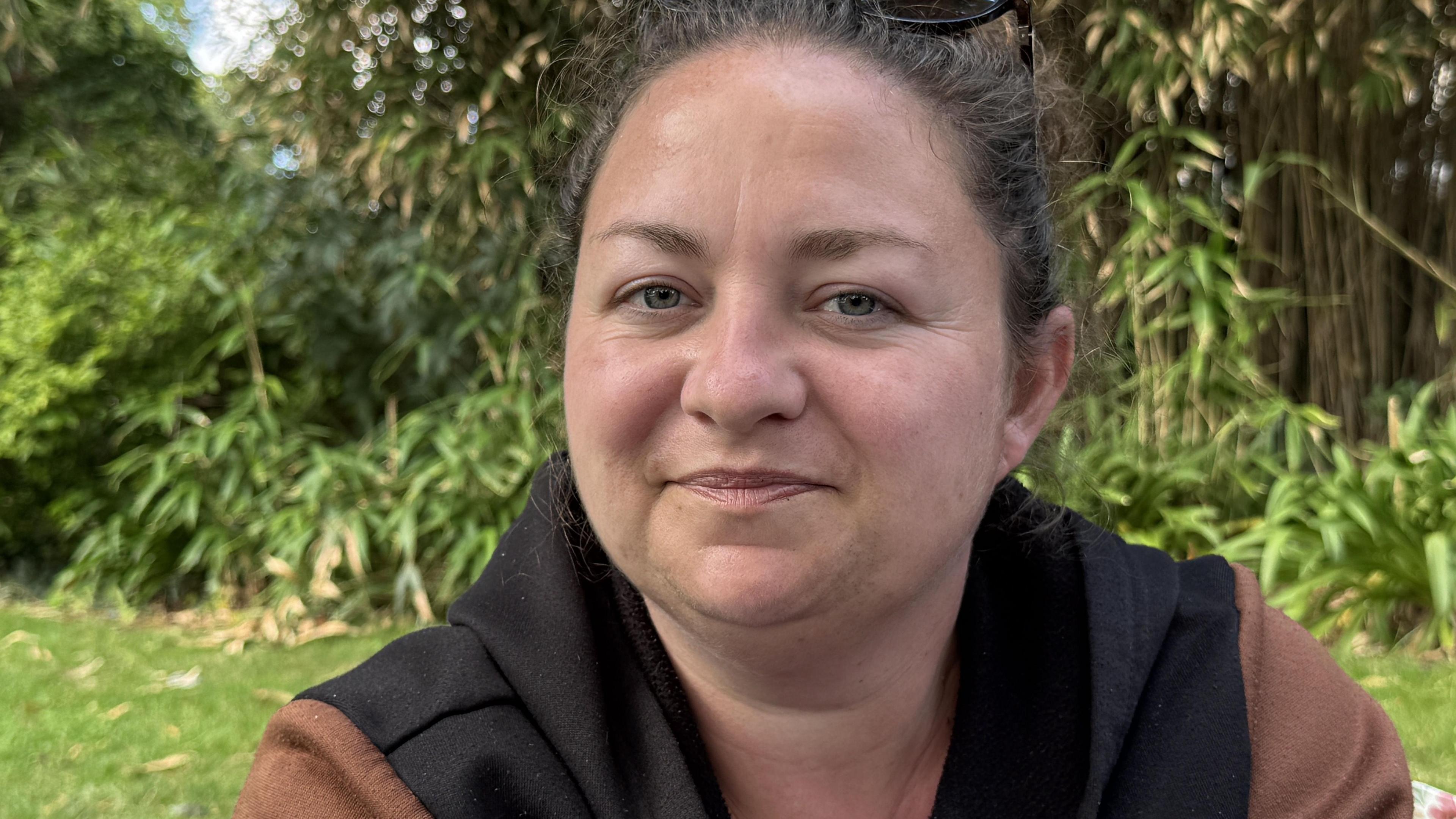 A woman sits in a park surrounded by bamboo stalks and thick foliage. She has dark hair, pulled back from her face, brown eyes and an olive complexion. She's wearing a brown top with a black gilet and she has sunglasses on top of her head.